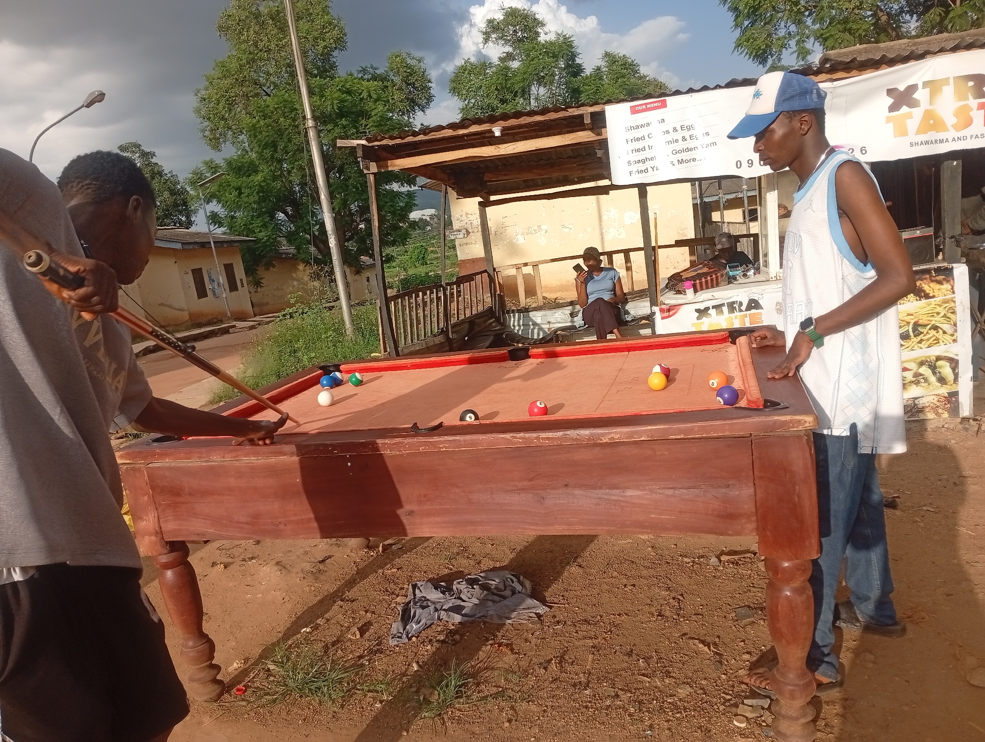 The image depicts an outdoor scene in Rafin Par, Nigeria, centered around a wooden pool table. Three individuals are visible. In the foreground left, a person wearing a grey t-shirt and black shorts is positioned behind the pool table, holding a pool cue. To the right, a person in a blue and white cap, a white and blue sleeveless jersey, and blue jeans stands beside the table, looking towards it. In the background, a person wearing a blue shirt is seated on a bench.
The pool table itself is made of dark wood and has a red felt surface. Several pool balls are scattered across the table, including solid colors like red, orange, and purple, as well as striped balls and a black 8-ball. A pool cue is resting on the table, and another is being held by the person on the left.
Behind the pool table, a small open-air structure with a corrugated metal roof provides shade. A sign on this structure lists menu items such as "Shawarma," "Fried Chicken & Eggs," "Spaghetti," and "Fried Yam." Adjacent to this structure, a larger sign displays "XTRA TASTE SHAWARMA AND FAST FOOD."
The environment appears to be a rural or semi-urban setting. The ground is covered in dirt and some sparse vegetation. Trees are visible in the background, and a cloudy sky suggests the time of day might be late afternoon or evening. Buildings with simple architecture are also present in the distance. The overall impression is a casual, open-air gathering space, likely a food stall or gathering point with recreational amenities.
