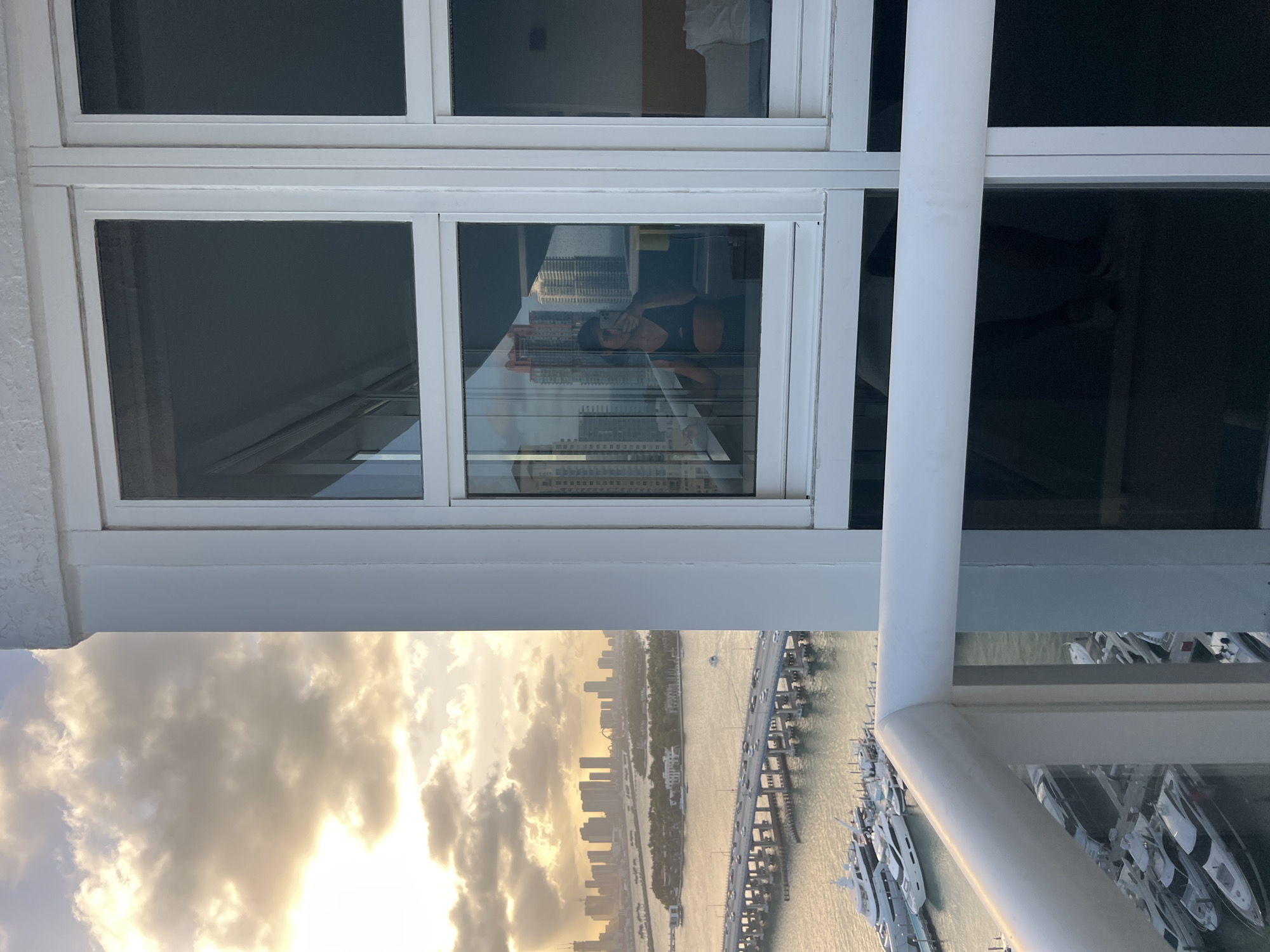The scene depicts a view from a high-rise building, possibly a condominium, in Miami Beach, United States. The camera is positioned looking out a multi-pane window and over a balcony railing. The visible view includes a cityscape partially obscured by clouds at sunset or sunrise, Biscayne Bay, a causeway with vehicular traffic, and a marina filled with yachts. The reflection of a person is visible within the window panes, holding a mobile device, presumably taking a photo. The window and balcony structure appear to be made of white metal or coated material.
