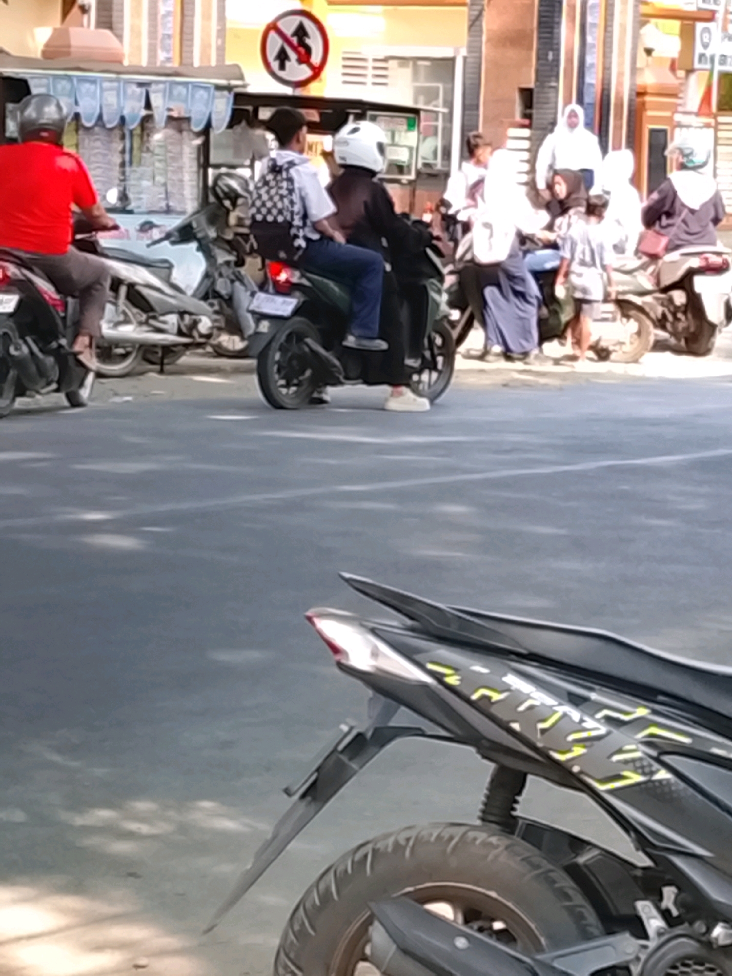 In Slawi, Indonesia, on a street, several people are on motorcycles. In the foreground, a person wearing a red shirt and helmet is on a black motorcycle. Behind them, another motorcycle carries two people. The passenger in the back wears a checkered backpack and a white helmet. Further in the background, a group of people on motorcycles are congregated, some wearing white headscarves. The environment suggests a sunny day, with shadows cast on the asphalt road. A traffic sign with red and white markings is visible in the upper left portion of the image.