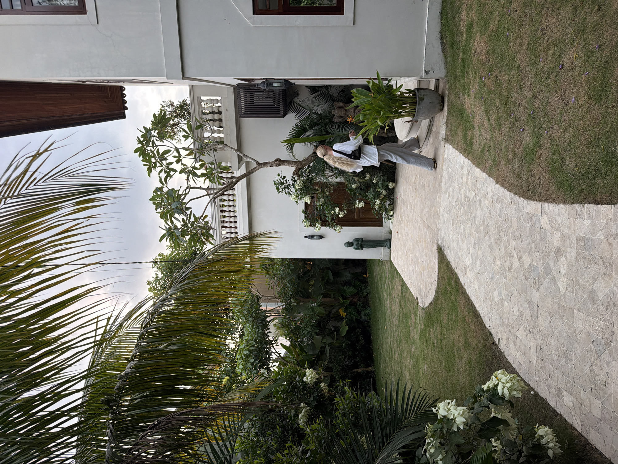 The image depicts a serene tropical garden and courtyard adjacent to white buildings, characteristic of North Kuta, Indonesia, under an overcast sky.
In the mid-ground, a person with long, blonde hair styled in a ponytail stands casually on a light-colored, irregularly shaped paved pathway. They are wearing a white long-sleeved shirt, light grey trousers, and a black cross-body bag. The person is turned slightly to their right, with their left arm raised, perhaps in a gestural pose or pointing, while their right arm is bent at the elbow.
The pathway curves around a manicured lawn dotted with small purple blossoms. Lush green foliage dominates the scene, including large palm fronds in the foreground, a prominent tree with white flowers (resembling frangipani) next to the person, and dense shrubs filling the areas between the path and the buildings. Behind the person, a decorative wooden lattice or panel is affixed to the white building wall. Further into the garden, a small, dark statue, possibly a deity like Ganesha, is nestled among the plants, with a potted plant beside it. A wall sconce is also visible on the building.
The architecture consists of clean white walls with brown-framed windows and decorative balustrades on upper levels, suggesting a resort or residential setting. The diffused lighting indicates an overcast day, providing soft shadows. No text is visible in the image.
