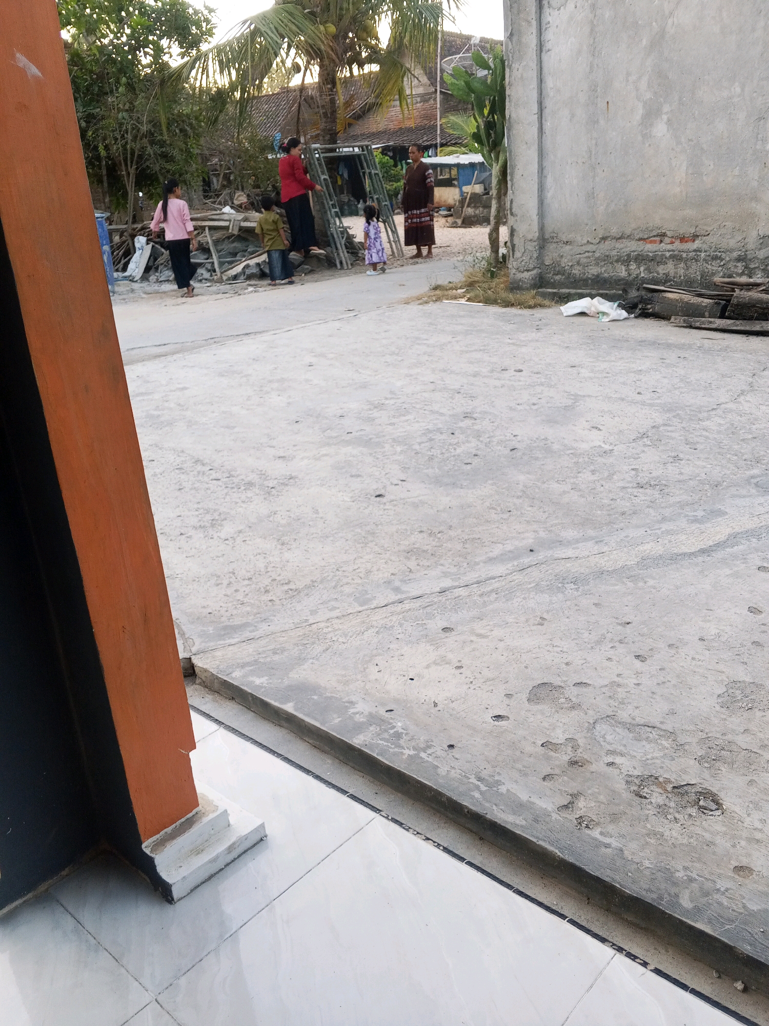 The image depicts a scene on a concrete ground, possibly a driveway or courtyard, in Gandrirojo, Indonesia. In the background, several individuals are visible. A person in a red top and black pants stands near some scaffolding and a pile of construction materials. To the left, two individuals, one in a pink top and black pants and another in a green top and dark pants, are walking. A young child in a purple dress is also present. Further to the right, another person in a brown patterned dress stands near a concrete wall. The environment suggests an outdoor setting with trees, a tiled floor area in the foreground, and a building structure to the right. The overall impression is one of a casual outdoor gathering or activity.