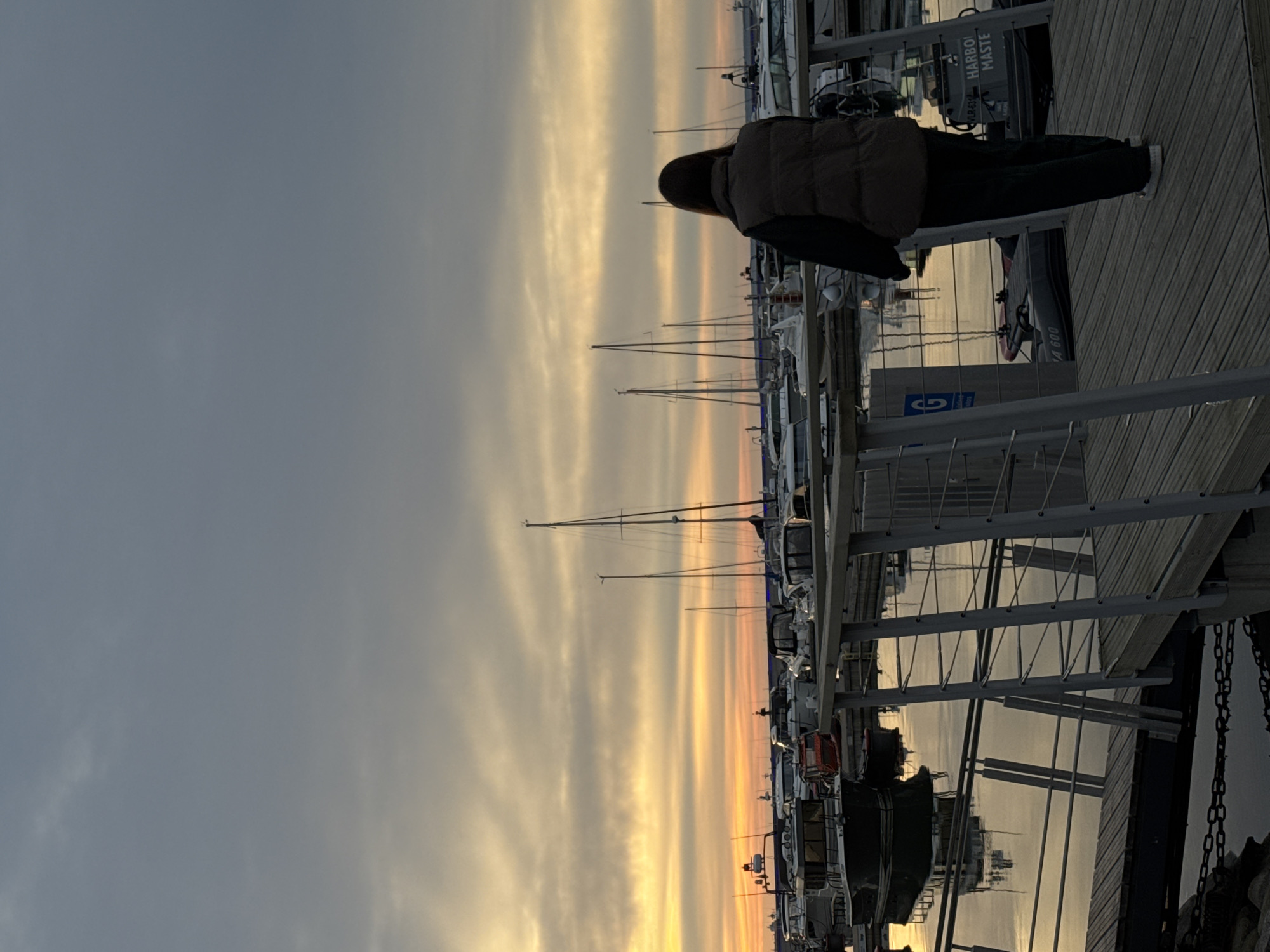 A person stands on a wooden pier at a marina in Tallinn, Estonia. The person, facing away from the camera, is wearing a dark puffer vest and dark pants. They are looking out at a body of water filled with various boats and yachts, many with their masts visible. The sky above is overcast with dramatic clouds, tinged with the orange and yellow hues of a sunset. The water reflects the colors of the sky and the boats. The pier has metal railings and wooden planks. In the background, there are signs on some of the boats. The overall atmosphere is peaceful and serene, with the setting sun casting a warm glow over the scene.