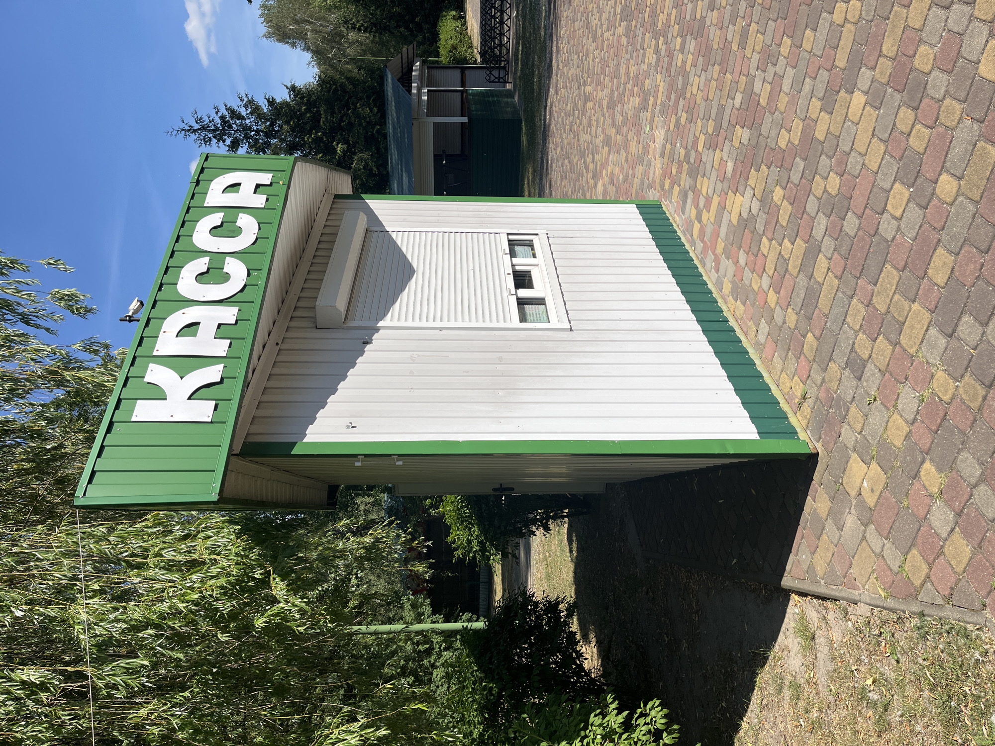An outdoor shot captures a small, white, booth-like structure with a green metal roof and base. The word "КАССА" (which translates to "cash desk" or "ticket office") is prominently displayed in large white letters on the green roof. The booth has a roll-down shutter covering a window opening. The ground in front of the booth is paved with colorful interlocking bricks. Lush green trees and foliage surround the structure, and the sky above is clear blue. Another, larger open-air booth with a green roof is visible in the background, to the right of the main booth. The scene appears to be a sunny day in a park or outdoor venue.