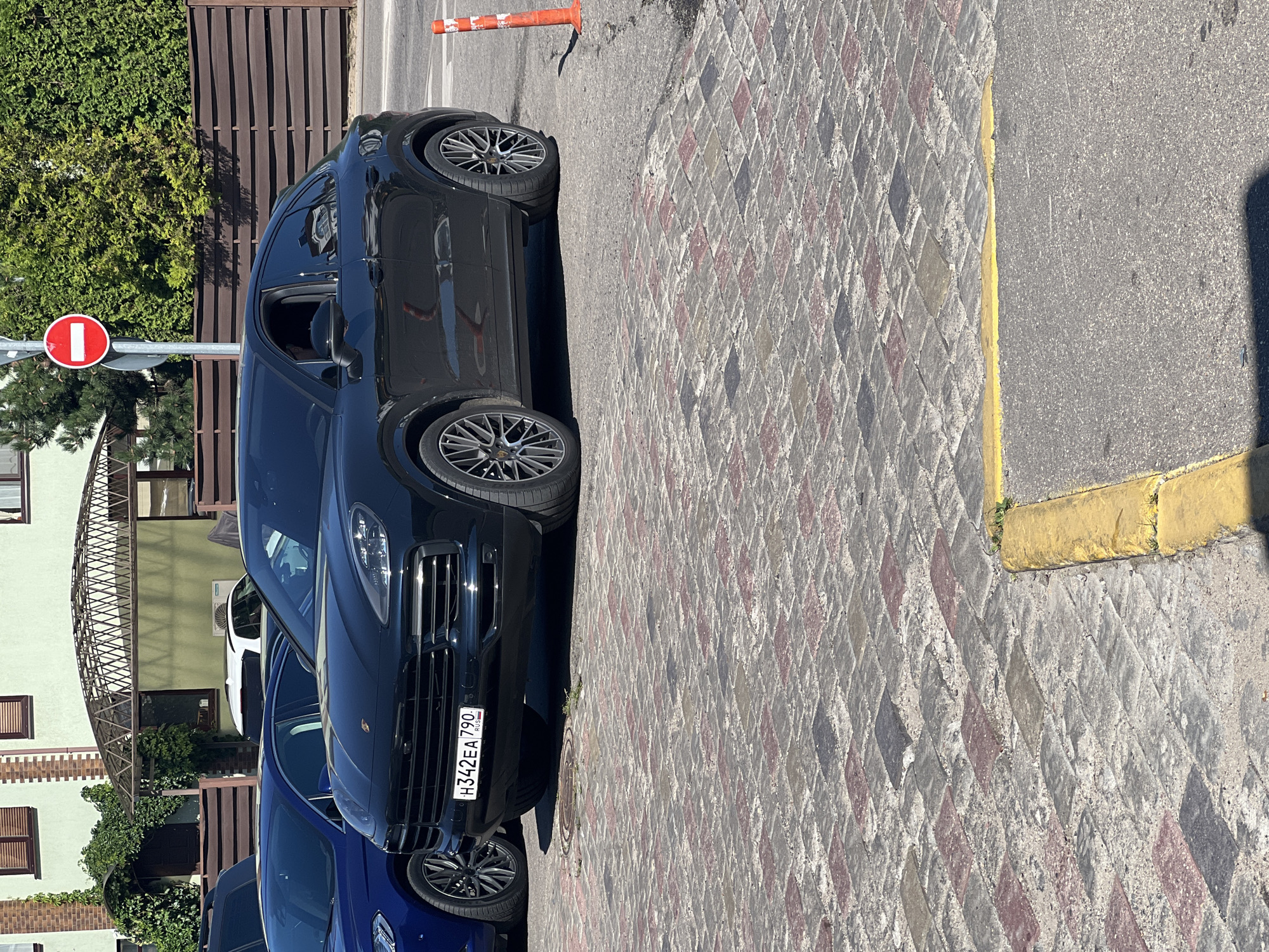 A black Porsche Cayenne Coupe is parked on a cobblestone surface next to a street. The car is facing left and has a license plate that reads "H342EA 790 RUS". In the background, there is a building with green siding and brown trim, a brown wooden fence, and lush green trees. A red and white circular sign is visible on a pole to the right of the car. Two orange traffic cones are also present on the right side of the image. The scene appears to be in a city.