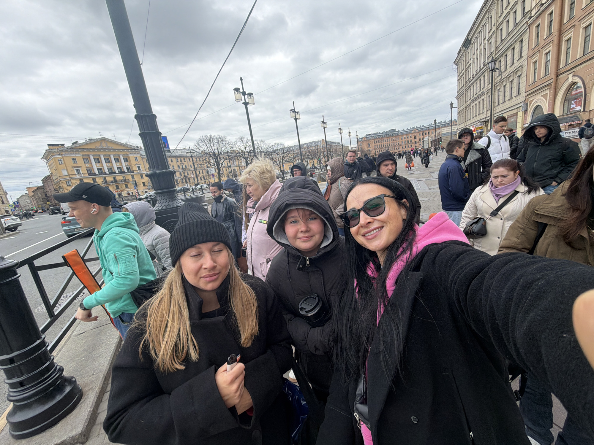 This bustling outdoor scene captures a selfie of three women in the foreground against the backdrop of a lively city street in Saint Petersburg, Russia, on an overcast day.
In the immediate foreground, a woman with long dark hair, black sunglasses, and a pink hoodie under a black coat holds the camera, smiling broadly. To her left, a younger individual with dark hair and a dark hooded jacket holds a black thermos, offering a slight smile. Further left, a woman with blonde hair, wearing a black beanie and a black coat, has her eyes closed or squinting, with a relaxed expression, holding a small object in her hand.
Behind them, the scene is crowded with numerous pedestrians, suggesting a popular area. People are dressed in various casual outerwear like coats, jackets, and hoodies, indicating cool weather. Cars are visible on the street to the left. The background is dominated by grand, multi-story buildings characteristic of European architecture. A prominent light yellow building with columns and decorative elements is visible to the left, standing out against the cloudy sky. Black street lamps and railings line the sidewalk.
The activity central to the image is the selfie being taken by the three women, conveying a casual and friendly moment. The overall impression is one of a busy urban environment, possibly a tourist area or a major thoroughfare. On a distant building to the far right, "Pro Max" is legible text. The sky is completely covered with gray clouds, suggesting it's daytime but without direct sunlight.