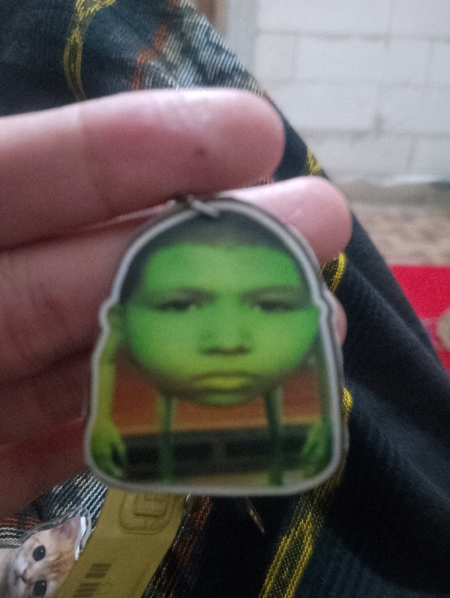 In Sowan Kidul, Indonesia, a person's finger is holding a green keychain with a cartoonish face printed on it. The background consists of a dark fabric with yellow stripes, and a hint of a red surface. Below the keychain, a small cat with light-colored fur and dark eyes is visible, lying on a yellow tag with some text. The image is taken from a close-up perspective, focusing on the keychain and the hand holding it.