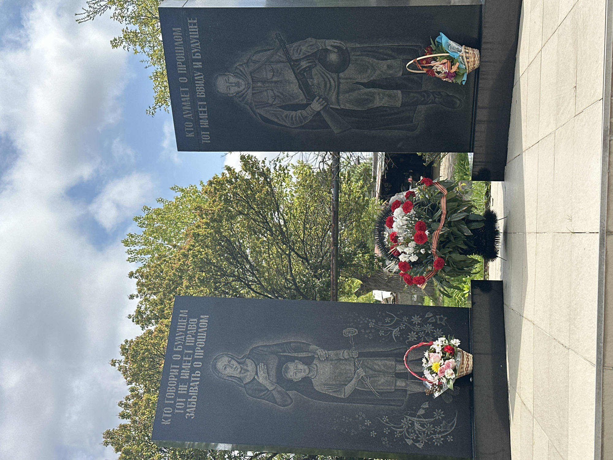 The image shows two adjacent black granite monuments adorned with etched images and floral tributes. The monument on the left depicts a woman with a child and flowers, while the one on the right portrays a soldier with a rifle. A basket of flowers sits near the base of the left monument, and a wreath of red and white flowers rests between the two. Both monuments are inscribed with text in Russian. The background includes a partially visible tree with green foliage against a blue sky with scattered clouds. The monuments are situated on a stone walkway, suggesting a memorial or cemetery setting. The location is Novomikhaylovsky, Russia.