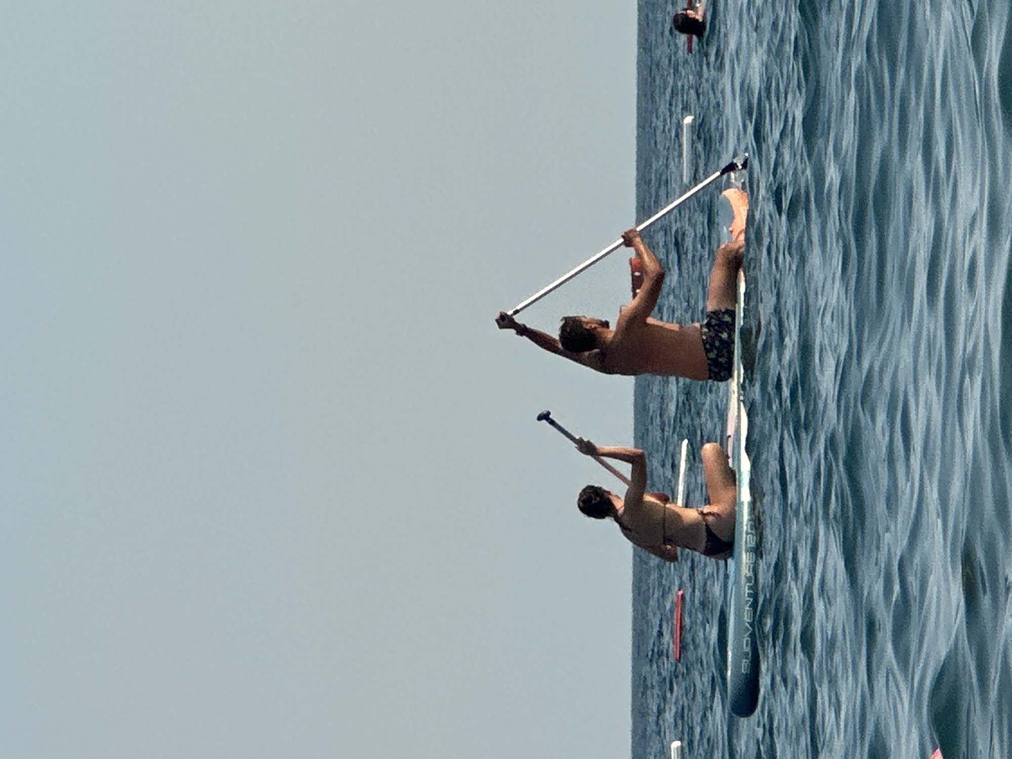 Two individuals are paddleboarding on a body of water, possibly the Adriatic Sea off the coast of Boreti, Montenegro. The person on the left, viewed from behind, is wearing a dark bikini and is seated on a paddleboard, holding a paddle with both hands, with the paddle head resting on the water. The person on the right, also viewed from behind, is wearing patterned swim trunks and is seated on a paddleboard, holding a paddle overhead with their right hand and the shaft of the paddle with their left hand, which is extended upwards. Their paddle is elevated above the water. Both individuals appear to be engaged in paddleboarding. In the background, the water is a deep blue, with moderate ripples. The sky is clear and a pale blue. There are other paddleboards visible in the distance, one with a red surface, and another person in the water. The overall scene depicts a recreational activity on a sunny day.