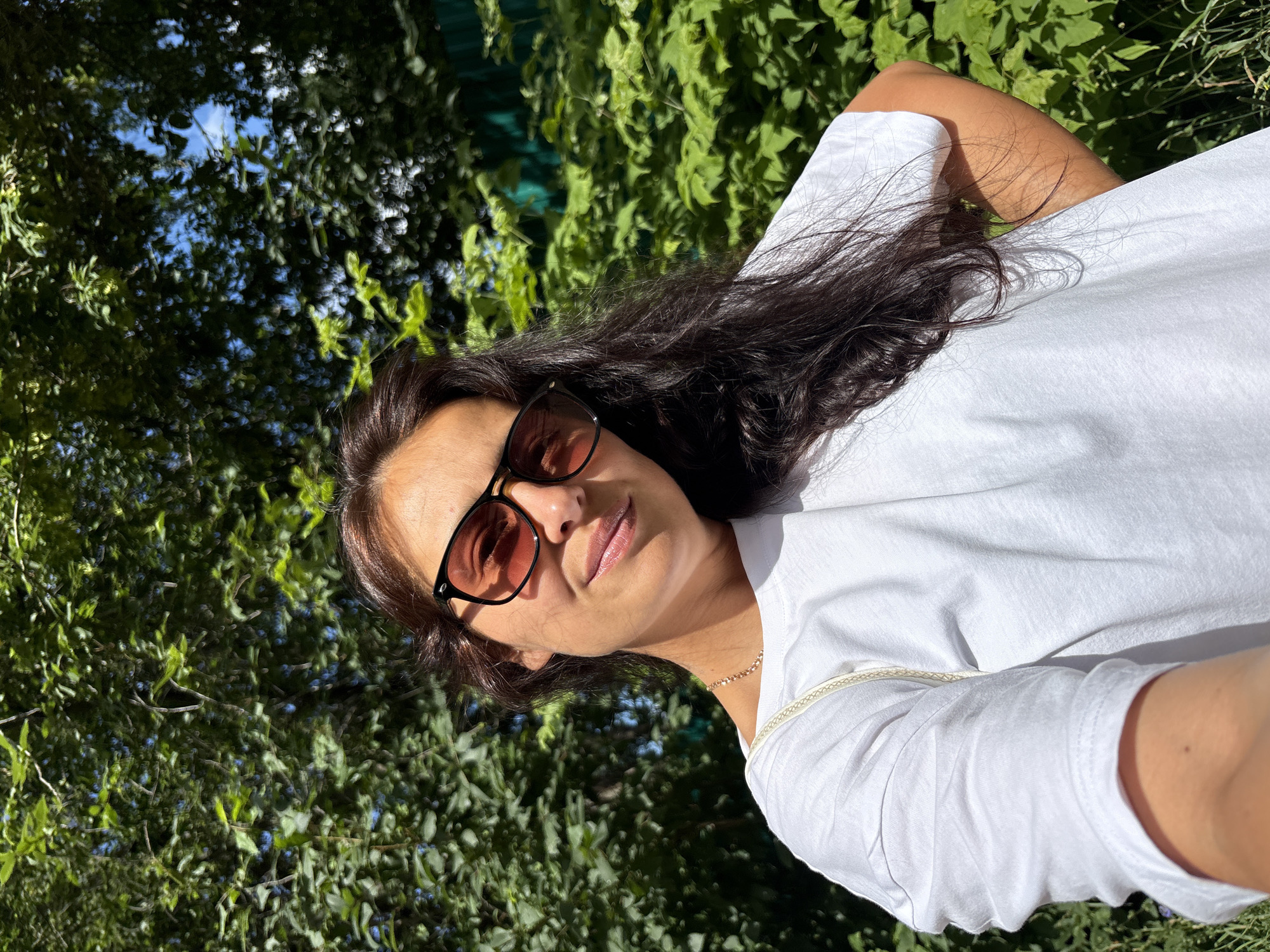 A woman is taking a selfie outdoors. She is wearing a white t-shirt and sunglasses. She has long, dark hair. The background is filled with green foliage and some blue sky visible through the leaves. The lighting suggests it is daytime and sunny. The specific location in the city of Chaikovsky, Russia cannot be determined from the visual information provided in the image.