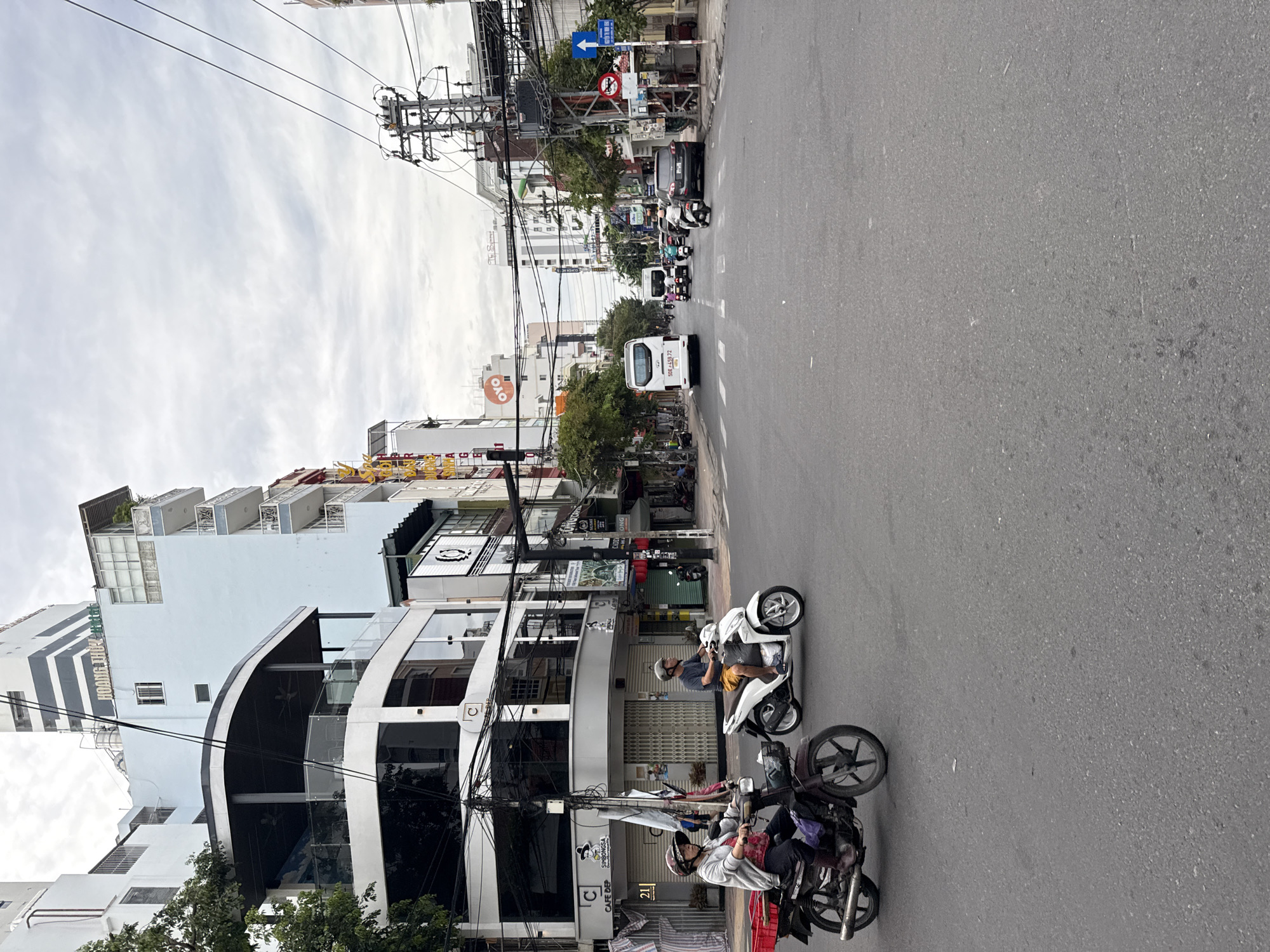 The image depicts a street scene in Nha Trang, Vietnam, likely a commercial district, under an overcast sky. The foreground shows a gray asphalt road with two individuals on motorcycles. The motorcycle on the left is a dark red, older model, ridden by a person wearing a gray jacket and a helmet, with a red basket attached to the rear. The motorcycle on the right is a white scooter, ridden by a person in a blue t-shirt and shorts, also wearing a helmet. They are traveling in the same direction.
The background is dominated by multi-story buildings of varying architectural styles. A prominent light blue building with white trim and balconies is visible on the left, bearing the sign "HOANG THUY". Adjacent to it are buildings with large glass windows and visible signage, including "CAFE DEP" and "Oyo". Further down the street, a white bus with the license plate "60E - 459.72" is seen, along with other vehicles, including cars and motorcycles. Numerous electrical wires and poles are a significant feature of the urban landscape, particularly on the right side of the street. Trees line parts of the sidewalk. Traffic signs, including a blue directional arrow and a red circular prohibition sign, are also present. The overall impression is one of a busy urban environment with a mix of modern and older architecture.