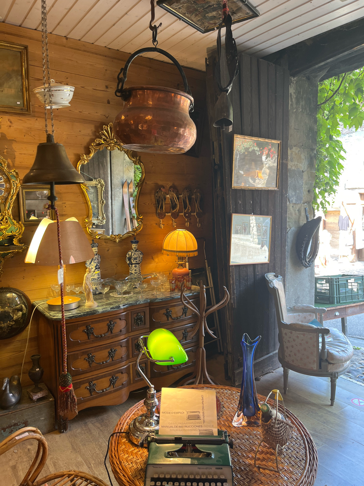 The image depicts an antique shop interior in Yvoire, France. The scene is filled with vintage items, including furniture, decorative objects, and lighting.
In the foreground, a round, woven table holds a vintage green typewriter, a blue glass vase, and a small wicker basket with a decorative bird. To the left, a wicker chair is partially visible.
Behind the table, a dark wood dresser with ornate metal hardware is laden with various items. These include a bright green desk lamp, glass vases, decorative figurines, and what appears to be an instruction manual.
Further to the left, a large ornate mirror with a gilded frame hangs on the wooden wall. A vintage bell lamp with a brown shade is suspended from the ceiling by a chain. Above it, a large copper pot hangs from a chain, and a smaller white bowl with a lid is also suspended.
To the right, a wooden coat rack stands near a dark wooden door frame. Beyond this, an antique upholstered armchair with a floral pattern is placed near a window or open doorway, through which greenery and a street scene are visible. Several framed pictures hang on the wooden wall. A tall, narrow wooden structure with horizontal slats is also visible.
The walls and ceiling of the shop are constructed from light-colored wood planks. The floor is also wooden. The overall impression is one of a cluttered but charming antique store displaying a variety of old items. No people are visible in the image.