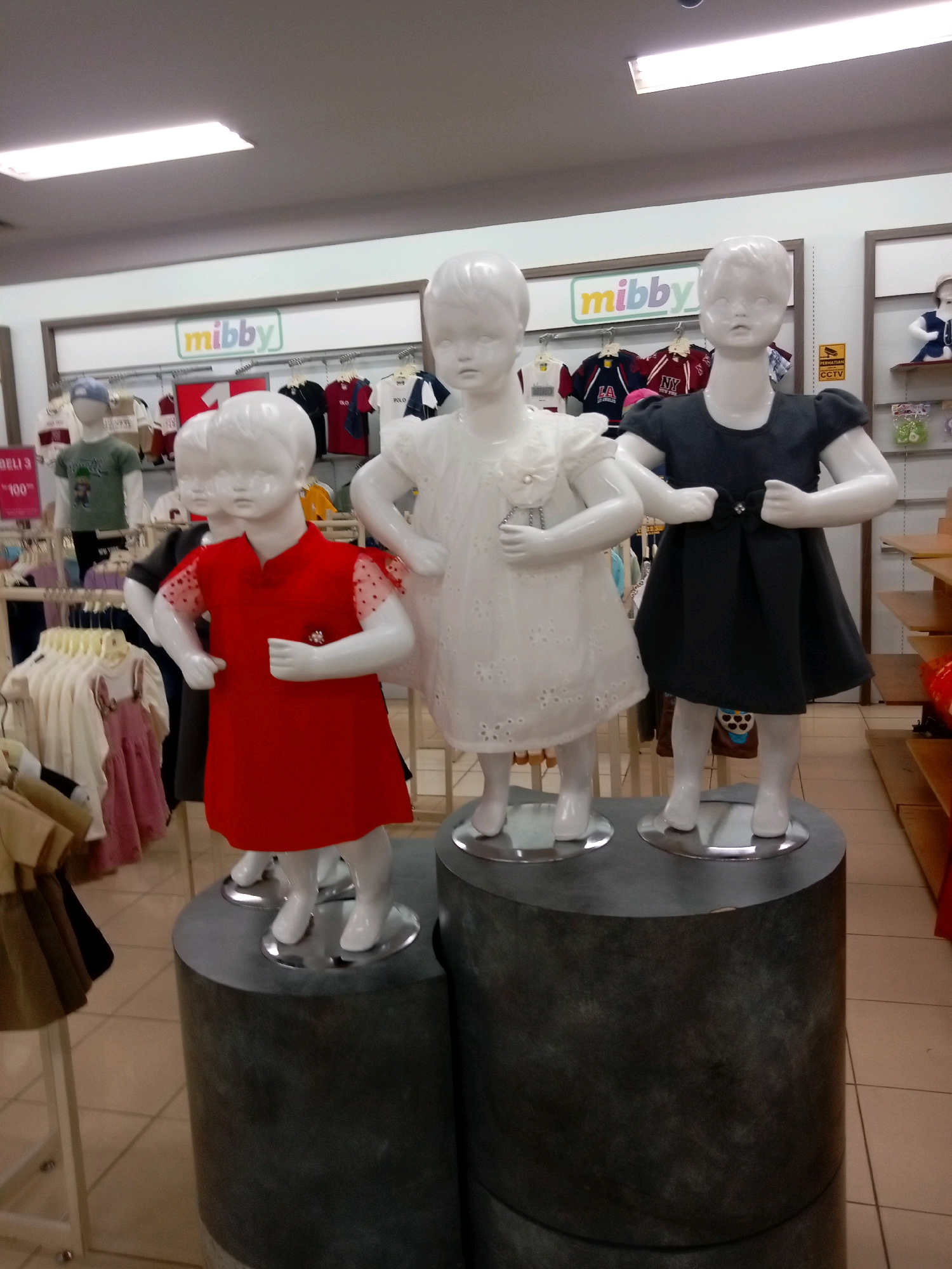 In a children's clothing store in Batam, Indonesia, three mannequins are displayed. The mannequins are positioned on cylindrical platforms and are dressed in children's clothing. The mannequin on the left is wearing a red dress, the middle mannequin is wearing a white dress, and the mannequin on the right is wearing a dark dress. In the background, racks of children's clothing are visible, along with signage. The store appears to be well-lit, with fluorescent lights on the ceiling. The overall environment suggests a retail setting dedicated to children's apparel.
