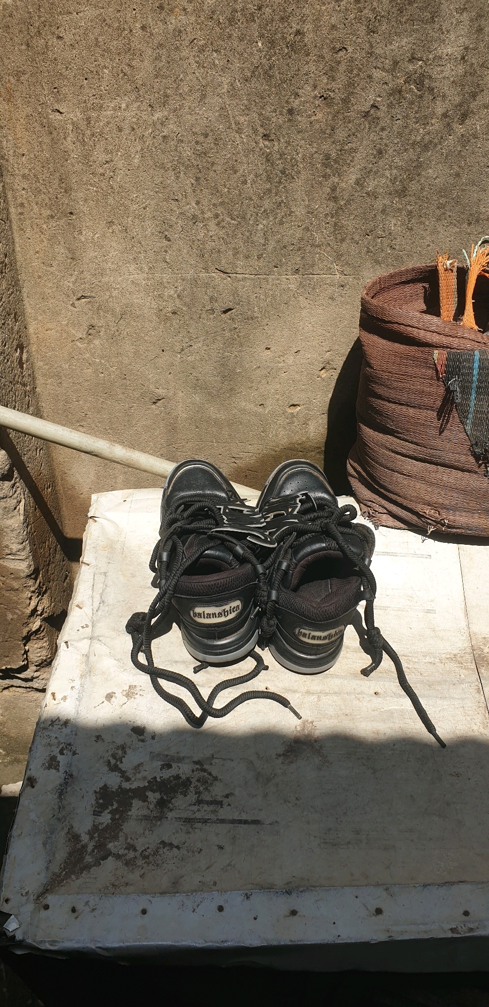 A pair of black athletic shoes, possibly a size 9 or 10, are chained together with black laces and placed on a worn, off-white surface. The shoes appear to be a men's style, with a bulky design and white accents on the sides. One shoe has the word "balan shies" visible on its heel. The surface they rest on is textured and shows signs of age and wear, with some dark staining. Behind the shoes, a large, rough-textured brown bag or sack is partially visible on the right side of the frame. The background is dominated by a textured, tan-colored wall, suggesting an outdoor setting. A shadow from an unseen object is cast across the shoes and the white surface. A white pipe runs horizontally in the background. The scene is located in Sabon Tasha, Nigeria.