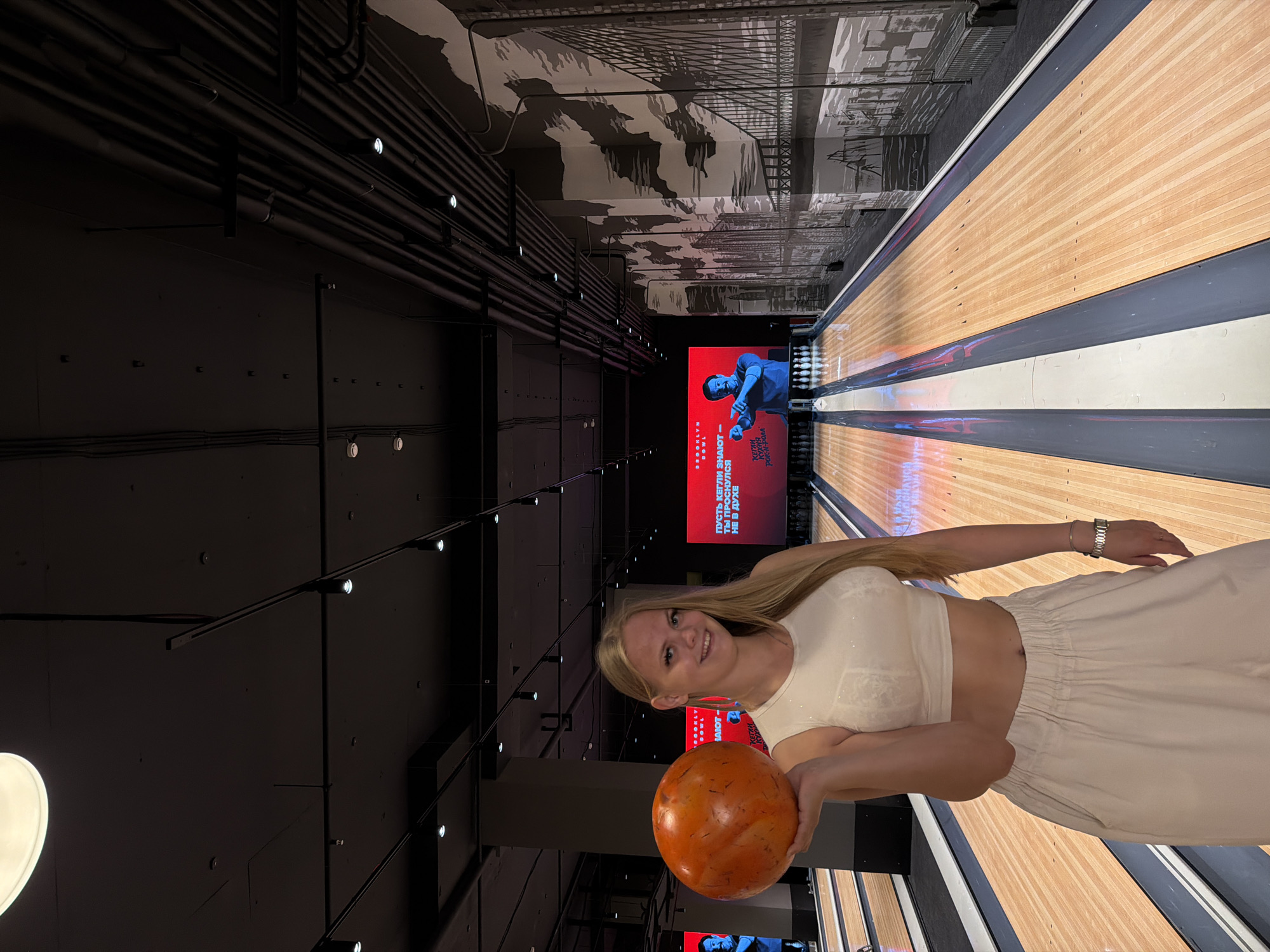 A young blonde woman is holding an orange bowling ball in her left hand, standing in a bowling alley. She is wearing a white crop top and loose, light-colored pants. The bowling alley has wooden lanes with dark gray gutters. A large digital screen behind her displays a man pointing and text in Russian, and to her right, a mural depicts a cityscape with bridges. The ceiling is dark with exposed pipes and lighting fixtures. The location is identified as "Brooklyn Bowl" in Izhevsk, Russia.