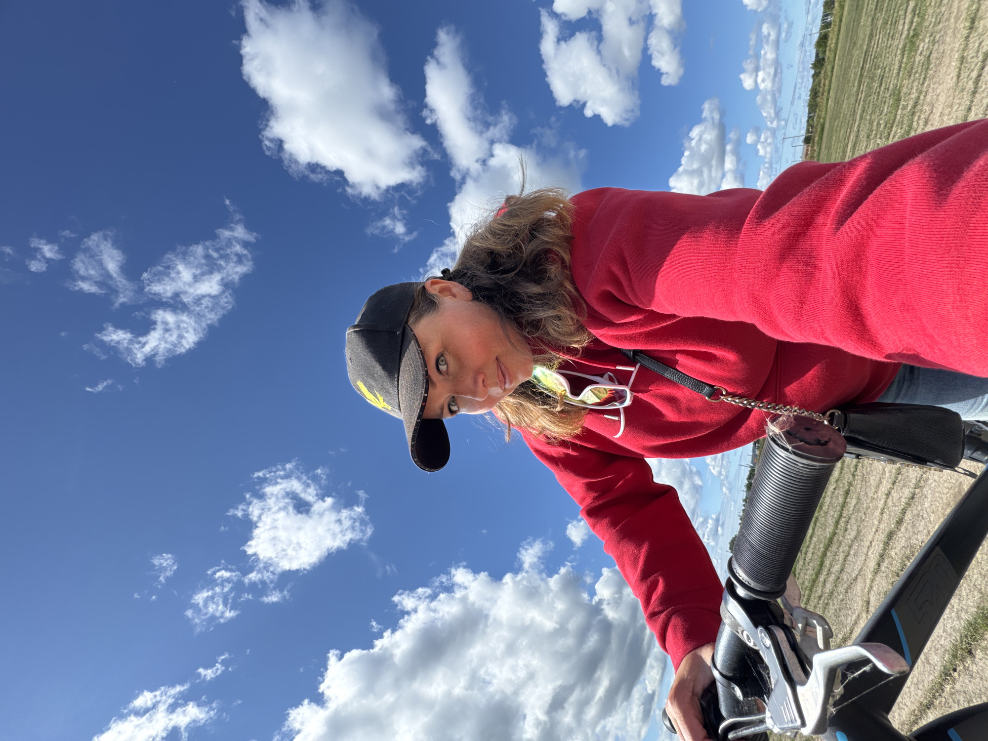 A medium shot captures a woman on a bicycle with a wide-angle perspective, looking directly at the camera. She is wearing a red hoodie and a black baseball cap with a yellow design. The background is a bright blue sky with scattered white clouds, and a field with parallel rows of dry grass. The bicycle handlebars and the woman's arm are in the foreground. The overall scene suggests an outdoor activity on a sunny day. The location is specified as Timmolovo, Russia.