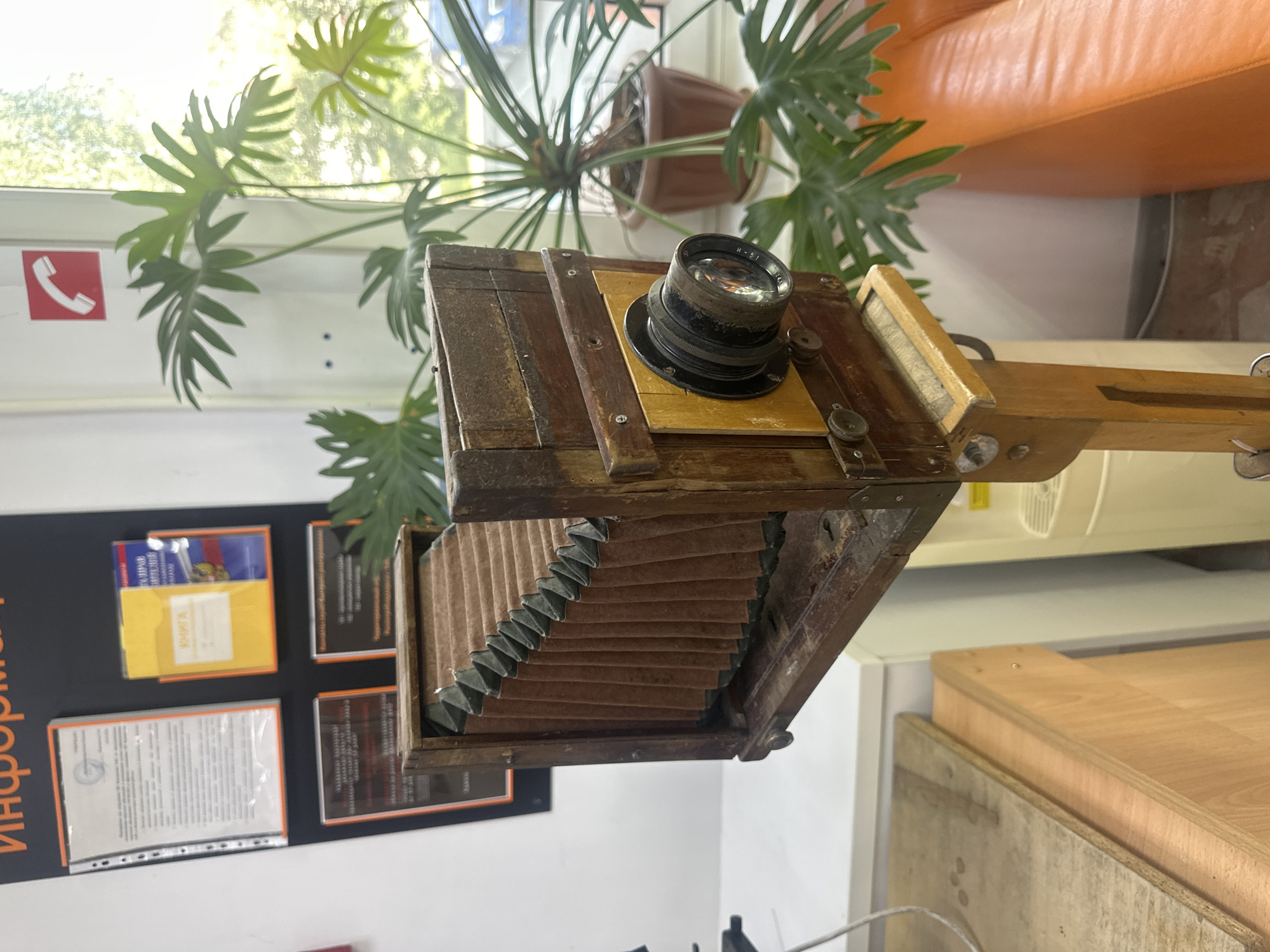 This is a medium shot, eye-level, indoor photograph of a vintage wooden bellows camera on a wooden stand. The camera is positioned slightly off-center to the right, facing left. The bellows are extended and made of brown material with green fabric lining. A large, round lens is attached to the front of the camera. The camera and stand appear aged, with visible wear and tear on the wood.
Behind the camera, on the left side of the frame, is a wall with several framed documents and posters. One poster at the top has orange text. To the right of the camera is a large potted plant with broad, green leaves, positioned near a window. Sunlight streams in through the window, creating a bright, slightly overexposed background with glimpses of trees and possibly a building outside. An orange sofa is visible in the lower right corner of the image.
The overall scene suggests a room that might be an office, museum exhibit, or storage area. The presence of informative posters and the vintage camera hint at a display or educational context.
Without further information or context, it is not possible to definitively determine the specific location in the city of Tobolsk, Russia, or provide any details about people, actions, or interactions, as none are present in the image.