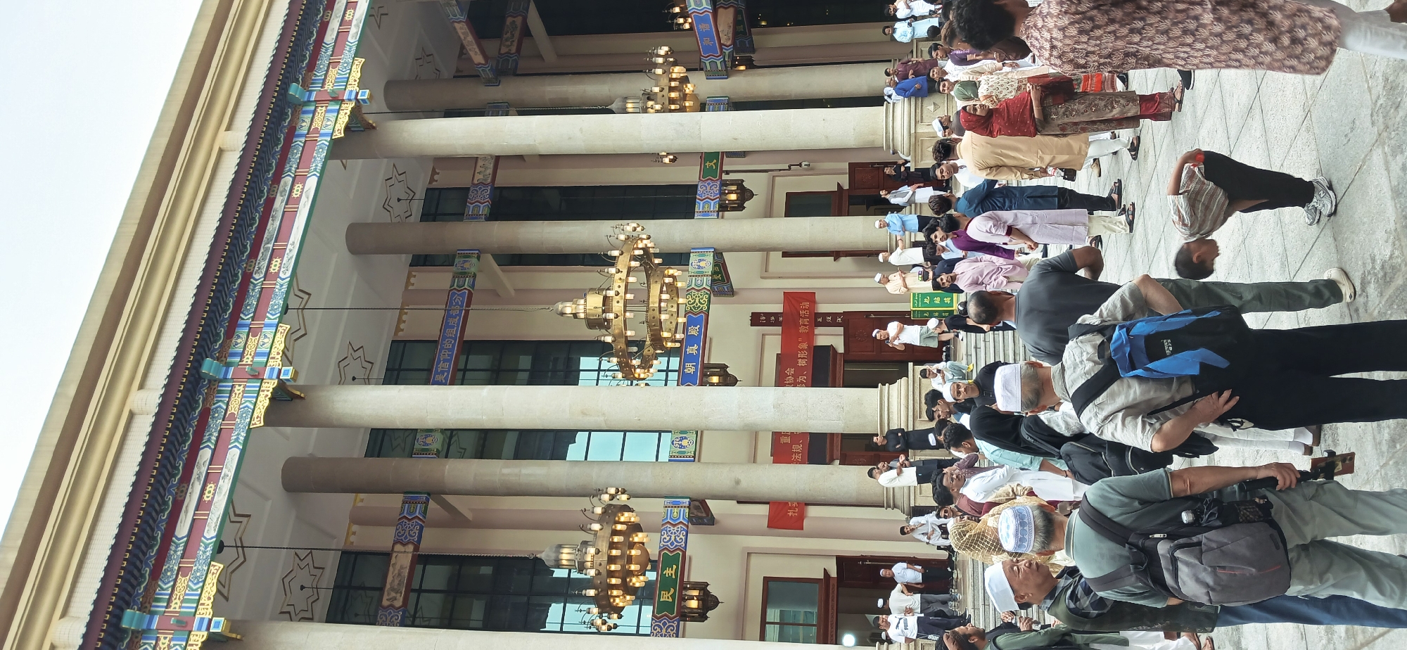 The image shows a group of Muslim men praying outside a building in Jiulongpo District, China. The building has a traditional Chinese architectural style, with a tiled roof and decorative columns. The men are kneeling and bowing in prayer. They are wearing a variety of clothing, including traditional Muslim dress and Western-style clothing. Some of the men have backpacks. There are banners written in Chinese characters on the walls of the building. The people are interacting in a religious manner. The environment seems to be an open space with reflective flooring, located outside a mosque or a similar Islamic building.