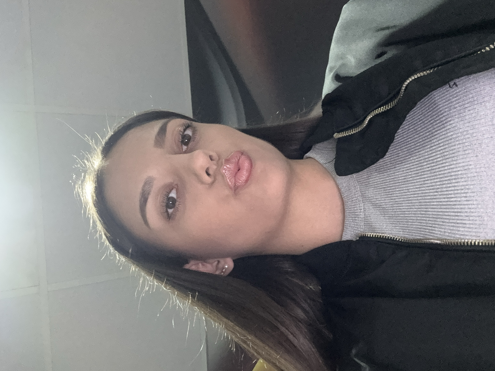 A woman is facing the camera, positioned with her head slightly tilted to the left. She has brown hair, brown eyes, and is making a kissing face. She is wearing a gray ribbed top and a black jacket with a silver zipper. The background is blurry and indistinct, but appears to include a light-colored wall or ceiling and a darker object that could be a vehicle interior. There is no identifiable information to suggest that the picture was taken in the city of Drenovets, Bulgaria.