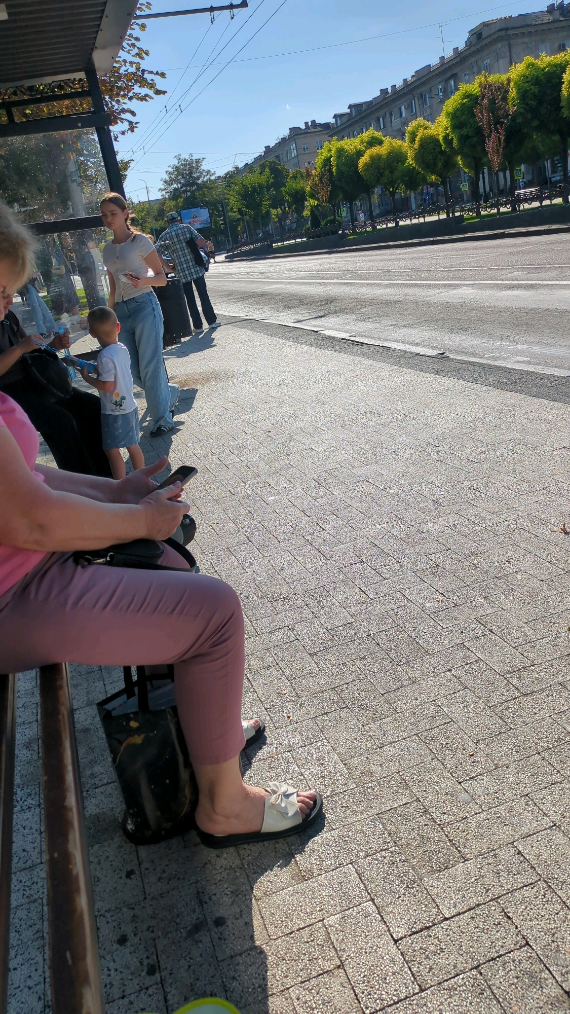 The image depicts a street scene in Chișinău, Moldova, likely near a bus stop, under a clear sky and bright sunlight. In the foreground, a woman is seated on a bench, wearing pink pants and white sandals with a bow detail. She is holding a smartphone in her hands. Adjacent to her on the bench is a black tote bag.
Further back, a young woman in a white t-shirt and jeans stands, also holding a phone. A young boy in a white t-shirt and blue shorts is beside her. Behind them, a man in a plaid shirt and dark pants is visible, carrying a bag.
The background features a street with traffic lanes, a sidewalk paved with textured rectangular tiles, and a row of trees lining the sidewalk. Buildings with classical architecture are visible in the distance. Overhead, power lines are strung across the sky, typical of urban infrastructure. The overall impression is a moment captured at a public waiting area in a city.