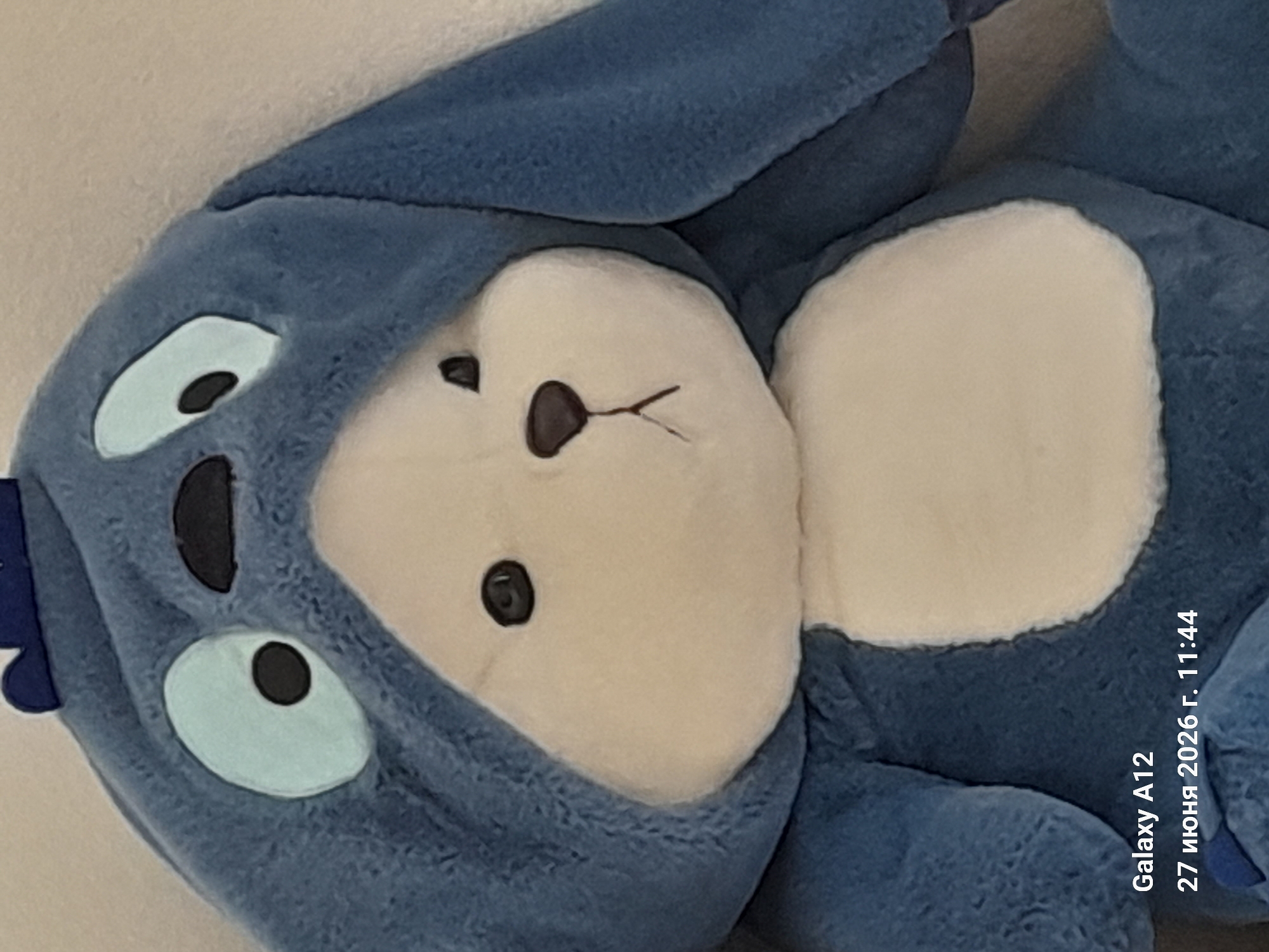 A close-up, eye-level, medium shot captures a blue plush toy with a white belly. The toy is wearing a blue hood with two large, light blue eyes, each with a black pupil. Above the eyes, on the hood, are two small black shapes that resemble eyebrows or part of the toy's character design. The toy has a white snout with a small black nose and a thin, dark line representing its mouth, which is slightly curved upwards. The background is a plain, light-colored surface. In the bottom left corner, text reads "Galaxy A12" and "27 июня 2026 г. 11:44," indicating the device and date/time of the photograph. The toy appears to be placed indoors.