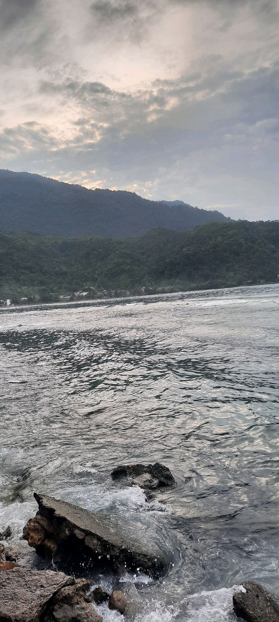 A vertical, low-angle shot captures a dramatic ocean view at Tapak Tuan, Indonesia. The foreground is dominated by dark, rugged rocks, some partially submerged in the choppy, gray water. White foam splashes against the rocks, indicating the movement of the waves. In the middle ground, the ocean extends towards a distant shoreline. The water is a shimmering expanse of silver and gray, with visible ripples and patterns formed by the waves. Along the distant shoreline, a dense, green hillside rises steeply, covered in lush vegetation. The hillside is a deep, dark green, with layers of mountains receding into the background. A faint scattering of buildings can be seen nestled along the base of the hillside. The sky above is overcast, with heavy, gray clouds that partially obscure the sun, casting a muted light over the entire scene. The overall impression is one of natural power and serene, yet imposing, coastal beauty.
