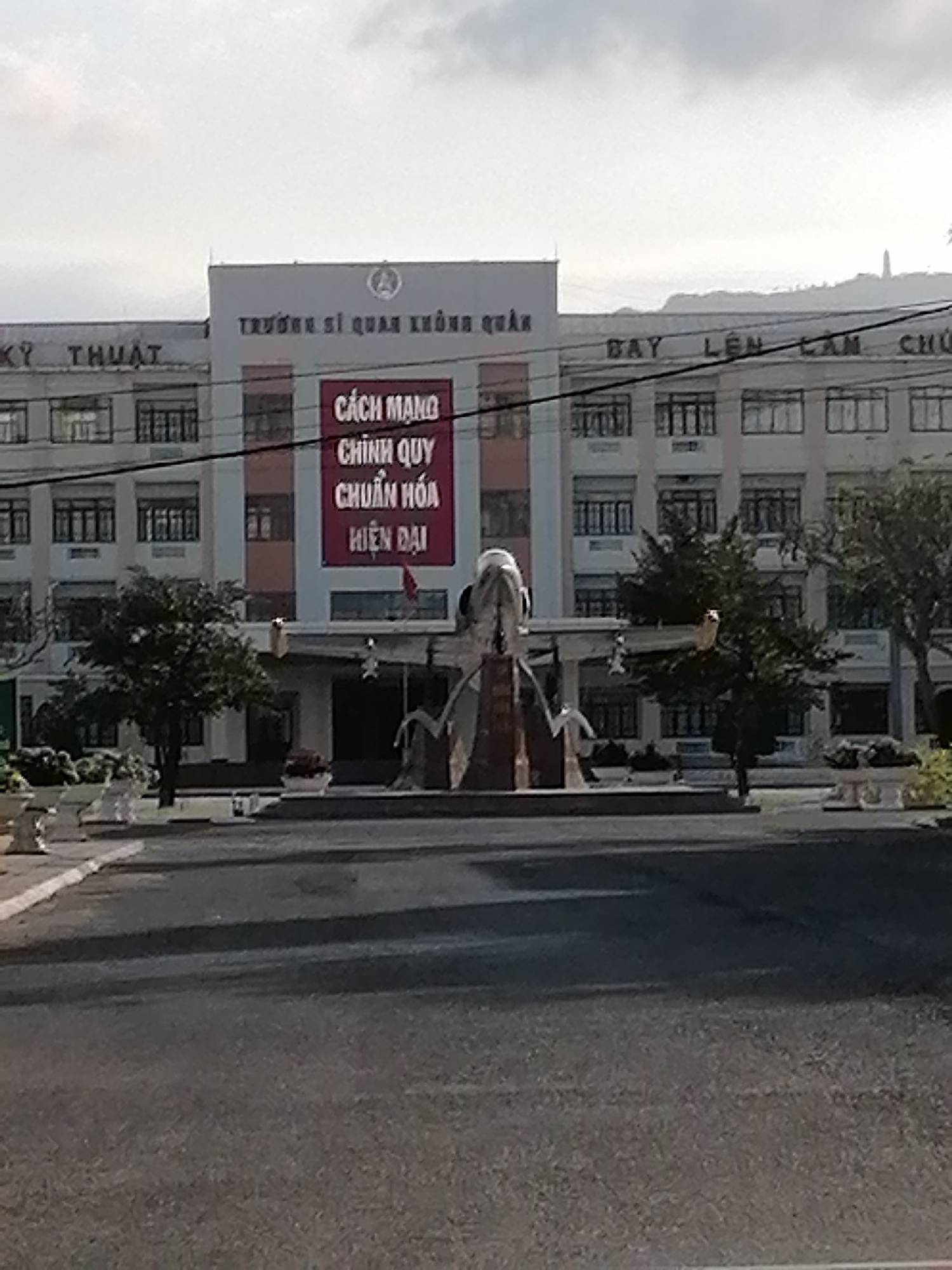 The image captures the view of a military building in Nha Trang Ward, Vietnam. The building is a large, multi-story structure with numerous windows and a sign in Vietnamese, "TRƯỜNG SĨ QUAN KHÔNG QUÂN" (Air Force Officer School). A large red banner is hanging on the building with Vietnamese text. In front of the building, there is a paved area with a sculpture, possibly of a military figure or symbol. Trees are planted on either side of the sculpture, and the road in front of the building leads towards the viewer. The sky is overcast.