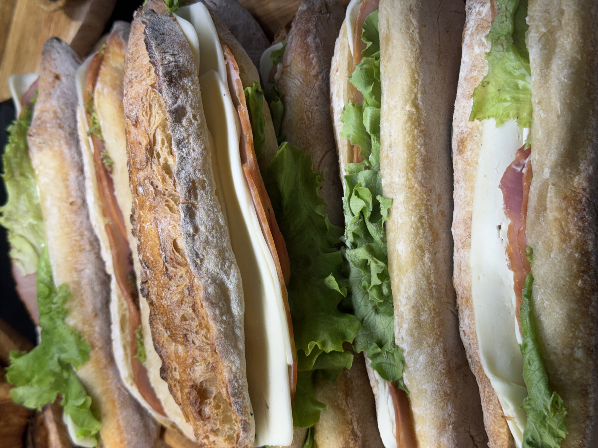 The image shows a close-up, overhead view of several sandwiches arranged next to each other. The sandwiches appear to be made with baguette-style bread, sliced lengthwise and filled with lettuce, cheese, and what looks like cured meat (ham or prosciutto). The bread has a crusty texture, and the fillings are fresh and green. The sandwiches are likely prepared and ready to be served.
There is no information in this image to suggest the location in the city of Tbilisi, Georgia.