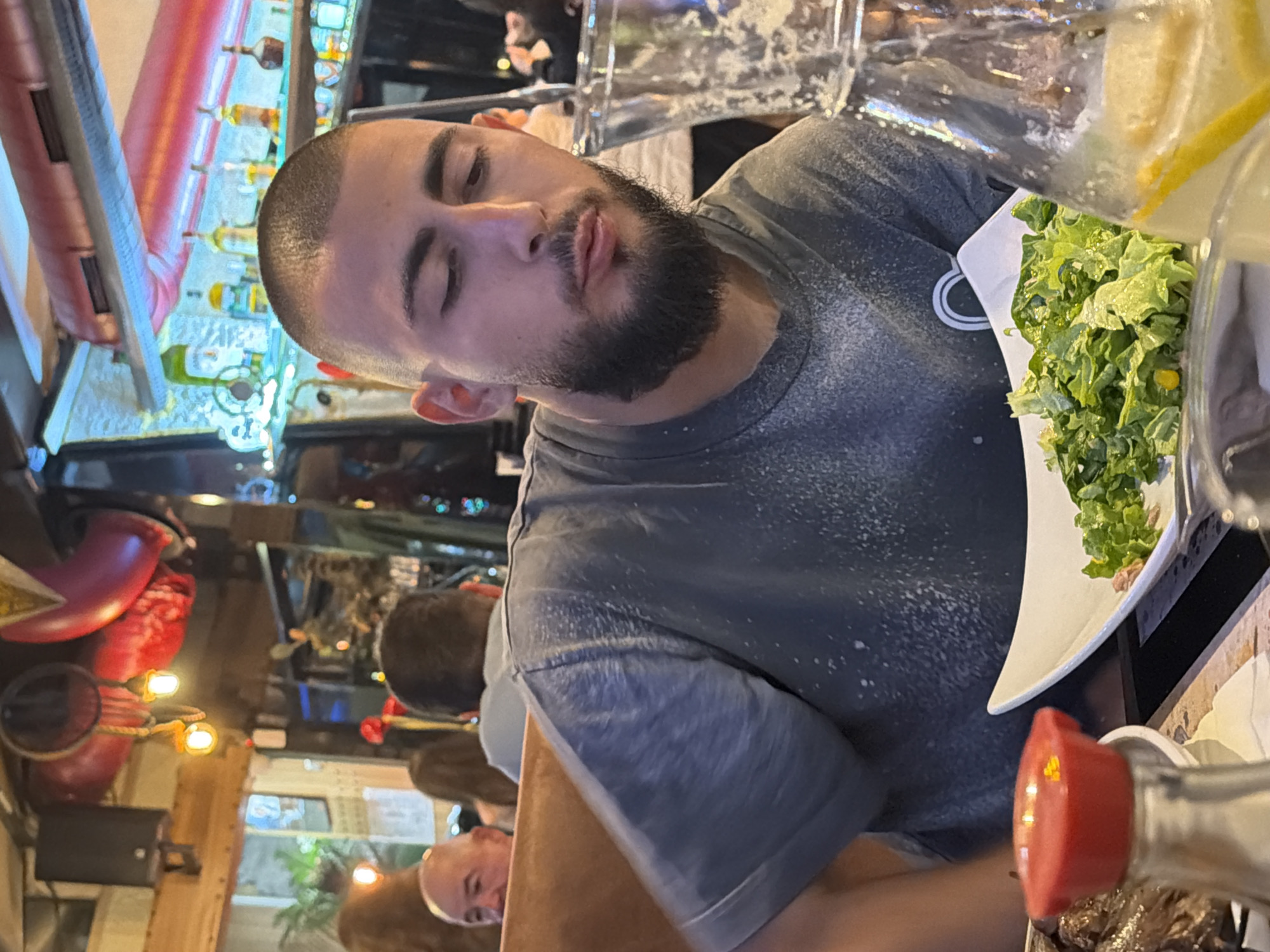 The image captures a man with a shaved head and a beard sitting at a table in a restaurant or bar. He is wearing a gray t-shirt and appears to be mid-action, possibly drinking or reacting to something.
In front of him is a plate of green salad, a glass of water with a lemon slice, and a container with a red lid. The table seems to be made of dark material, possibly wood.
The background is filled with other people, suggesting a moderately crowded environment. Various elements typical of a bar or restaurant setting are visible, including shelves with bottles and decorative lights. A red-colored decorative object hangs from the ceiling.
The location is noted as Unknown, Unknown, providing no specific geographic context.
