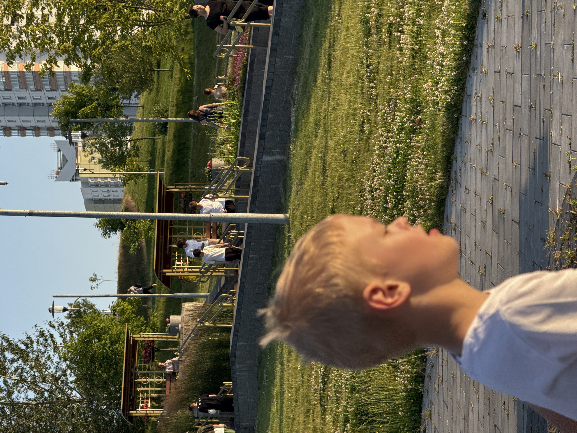 In an outdoor urban park setting, a young boy with blond hair is in the foreground, facing right in profile, wearing a white t-shirt. Beyond him, a paved walkway leads towards a grassy hill with scattered white wildflowers. Several people are visible on tiered walkways and near structures with pergolas. On the left, a person in a black t-shirt with white stars walks up stairs. A woman sits on a bench under a pergola on the left. Further up the stairs and to the right, two individuals in white shirts are seen, one standing and one walking. A woman with a backpack and a child are on the stairs in the middle ground. On the far right, a woman in a black dress stands on the stairs. In the background, apartment buildings rise above a treeline. The lighting suggests it is late afternoon or early evening.