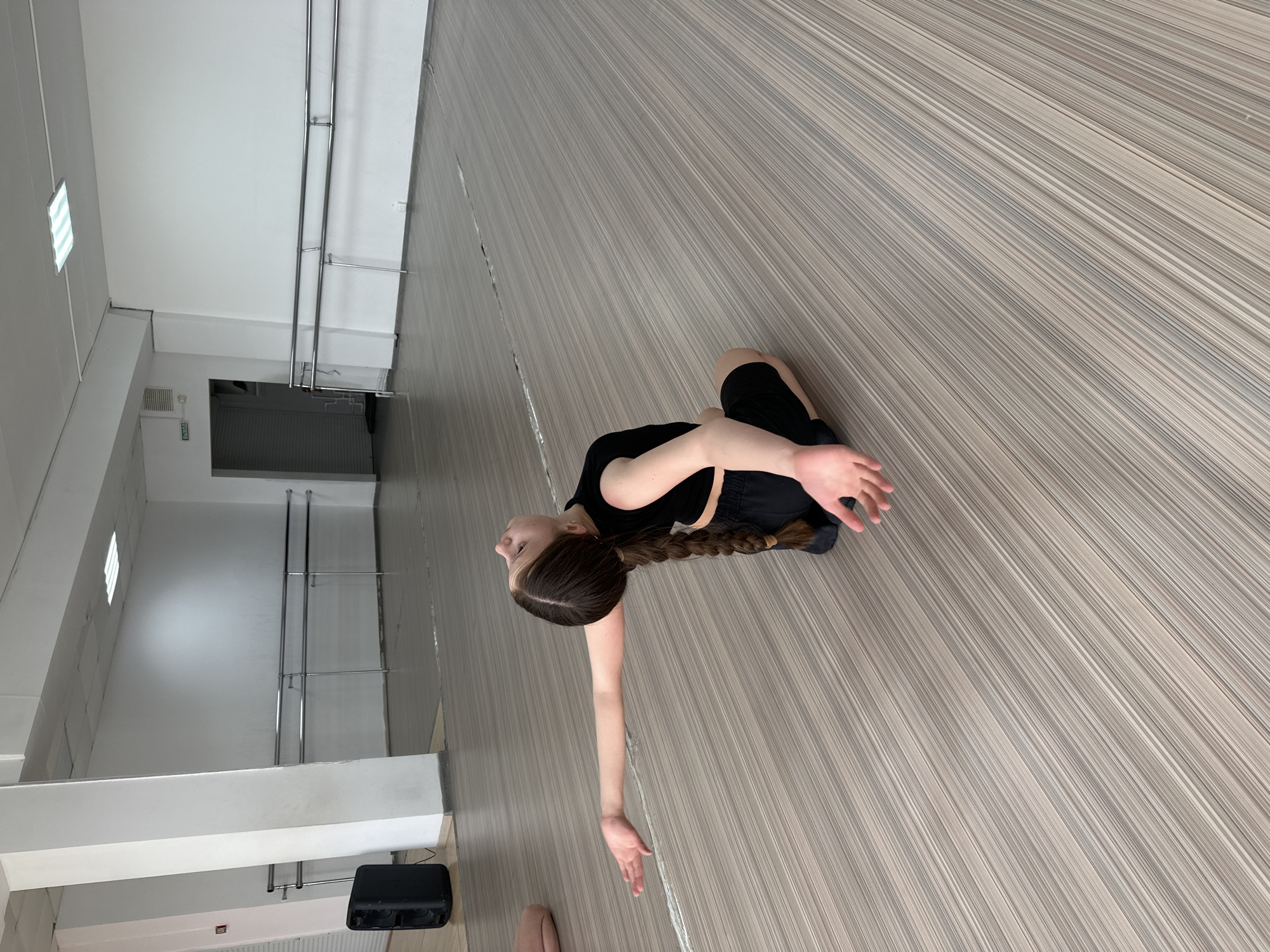 A young person is kneeling on a striped floor inside a building, likely a dance or exercise studio in Votkinsk, Russia. The person is wearing dark clothing, with long, braided hair. One arm is extended towards the lower portion of the frame, while the other arm is bent across their chest. The studio contains ballet barres along the wall, with a dark speaker or amplifier visible in the corner. The lighting is provided by fluorescent fixtures.