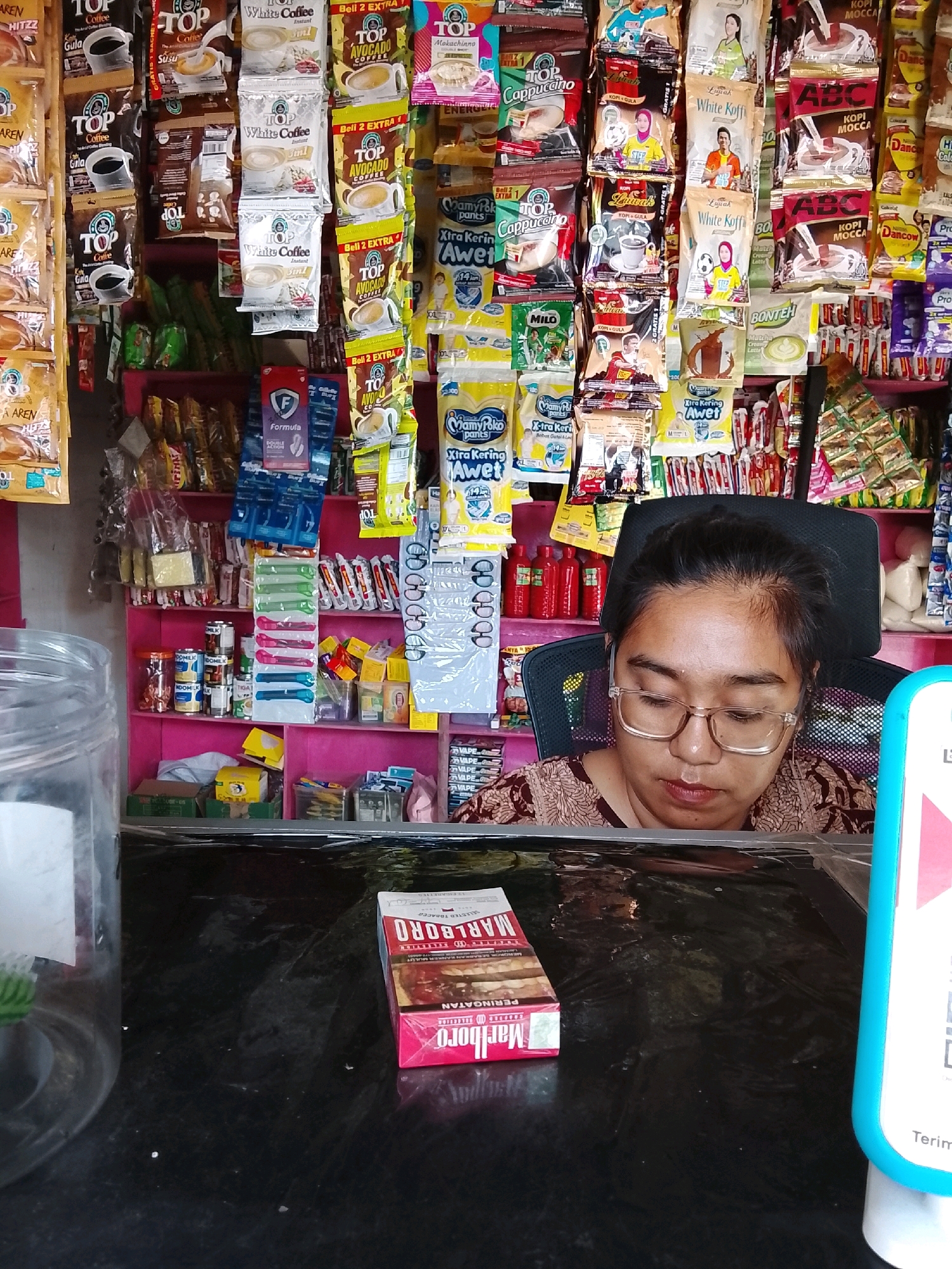 The image captures a close-up of a young woman with glasses, seated in front of a shelf stocked with various packaged food and beverage items. She is wearing a patterned shirt and appears to be looking down. In the foreground, a pack of Marlboro cigarettes rests on a dark, reflective surface, which is likely a counter or table. Behind the woman, the shelves are densely packed with colorful packaging, including coffee, tea, milk, and other processed food items. The overall setting suggests a small convenience store or warung. The presence of the woman and the goods on display indicate a retail environment. The image does not contain any overt actions or interactions, focusing instead on the static arrangement of the scene and the woman's presence within it.