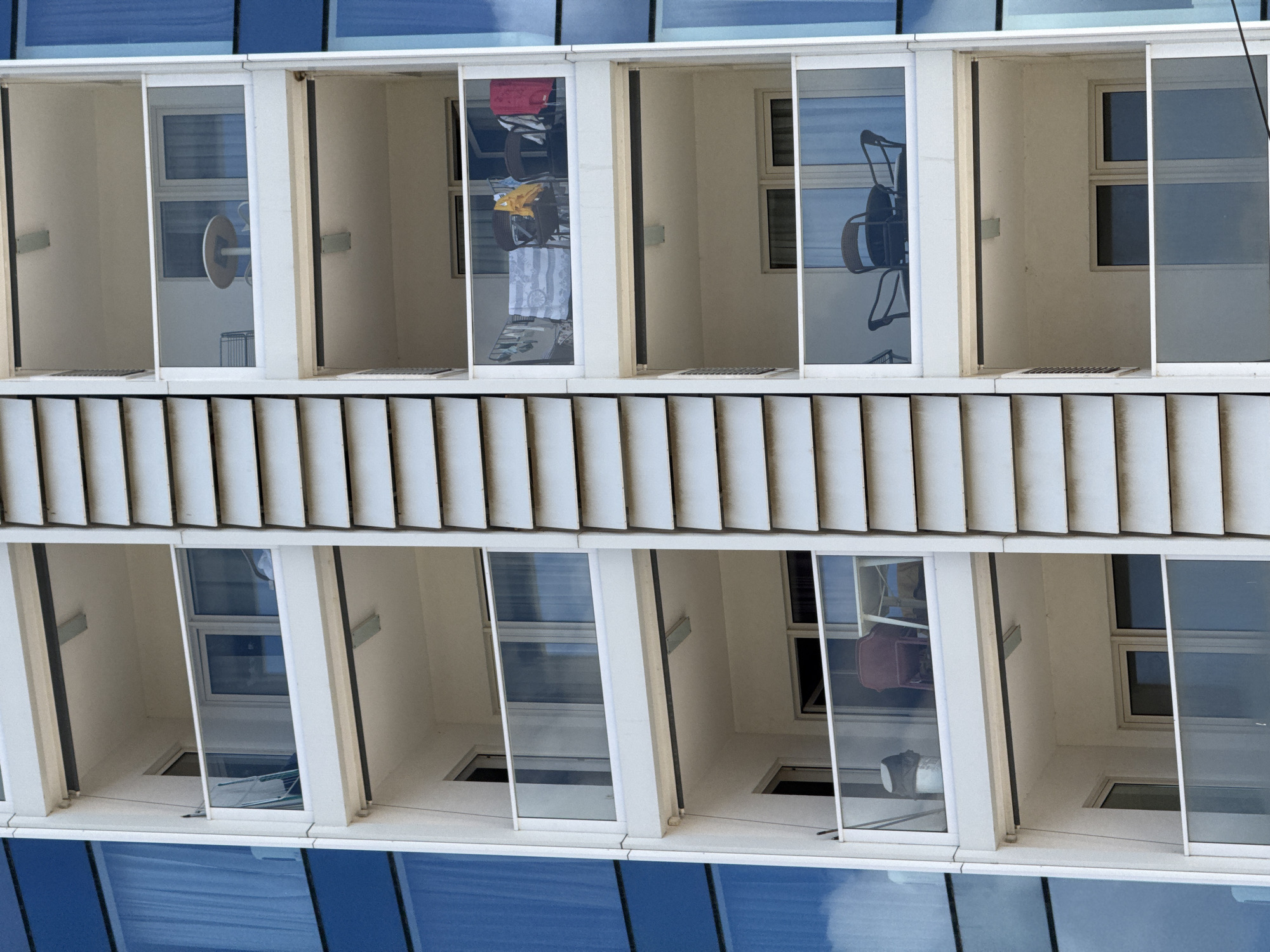 The image shows a close-up, high-angle view of a modern apartment building's exterior. The facade is characterized by a grid of balconies and windows. The balconies are narrow and enclosed with glass sliding doors, revealing the interiors. The building appears to be made of concrete and glass, with a ribbed metal overhang or roof structure at the top.
In several balconies, various objects are visible, suggesting occupancy. One balcony displays laundry hanging on a rack, including a red bag and towels. Another balcony shows a chair with a distinctive woven pattern, possibly an outdoor furniture piece. Reflections in the glass doors add to the scene, showing glimpses of the sky and possibly the opposite side of the building. The interiors of the balconies are sparsely furnished, with a window visible in each. The overall impression is one of residential living spaces in an urban or semi-urban environment. The time of day appears to be daytime, given the bright lighting and clear reflections. There are no visible people. The location context is Budva, Montenegro.