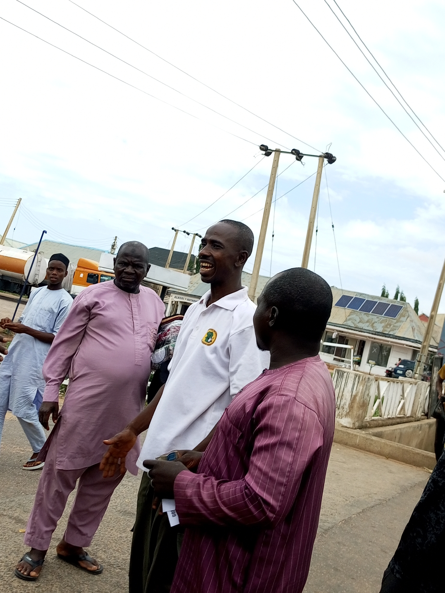 This media file shows a group of four men gathered on a paved surface, likely a street or sidewalk, in what appears to be an outdoor setting. The sky is overcast, suggesting either cloudy weather or a time of day with diffused light, possibly mid-morning or late afternoon. In the background, several objects are visible: utility poles with wires crisscross the sky, a large white and orange truck is parked, and a building with solar panels on its roof can be seen. Further in the distance, there are more structures, including a building with the partial text "ALMERI RESTAURANT".
The men are dressed in traditional attire. Two of them are wearing light-colored, loose-fitting garments, one in a pale blue and the other in a dusty rose pink. Another man is in a white polo shirt with a circular emblem on the chest. The fourth man is wearing a dark purple, pinstriped robe. The interaction between the men appears friendly and casual, with one man in the white shirt laughing heartily. One man in the pink robe is extending his hand, palm up, as if in greeting or explanation. Another man is holding some papers in his hands, one of which has partially visible text that includes "109". The overall impression is of an informal social gathering or a moment of discussion between individuals. The presence of the truck and the building with solar panels suggest a semi-urban or commercial area.