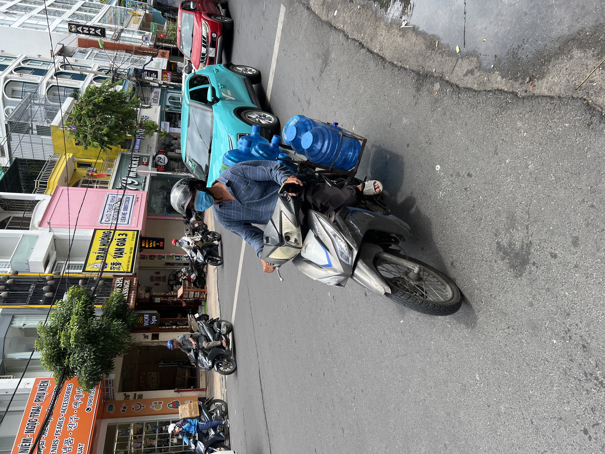 A man wearing a blue patterned shirt, dark pants, white sandals, a gray helmet, and a light blue face mask rides a gray Honda scooter. He is transporting five large blue water jugs, secured to a metal rack behind the seat. The scooter has a silver and blue front panel. The man is on a paved city street, appearing to be in motion. To his left, a turquoise car is driving in the same direction. To the right of the turquoise car, a red SUV is also driving. In the background, there are multi-story buildings with various signs, some in Vietnamese. One prominent sign is orange and says "NIEM - NGỌC TRAI - PHU KIỆN" and another sign says "WOW Spa". The overall scene depicts a bustling street in Nha Trang, Vietnam.