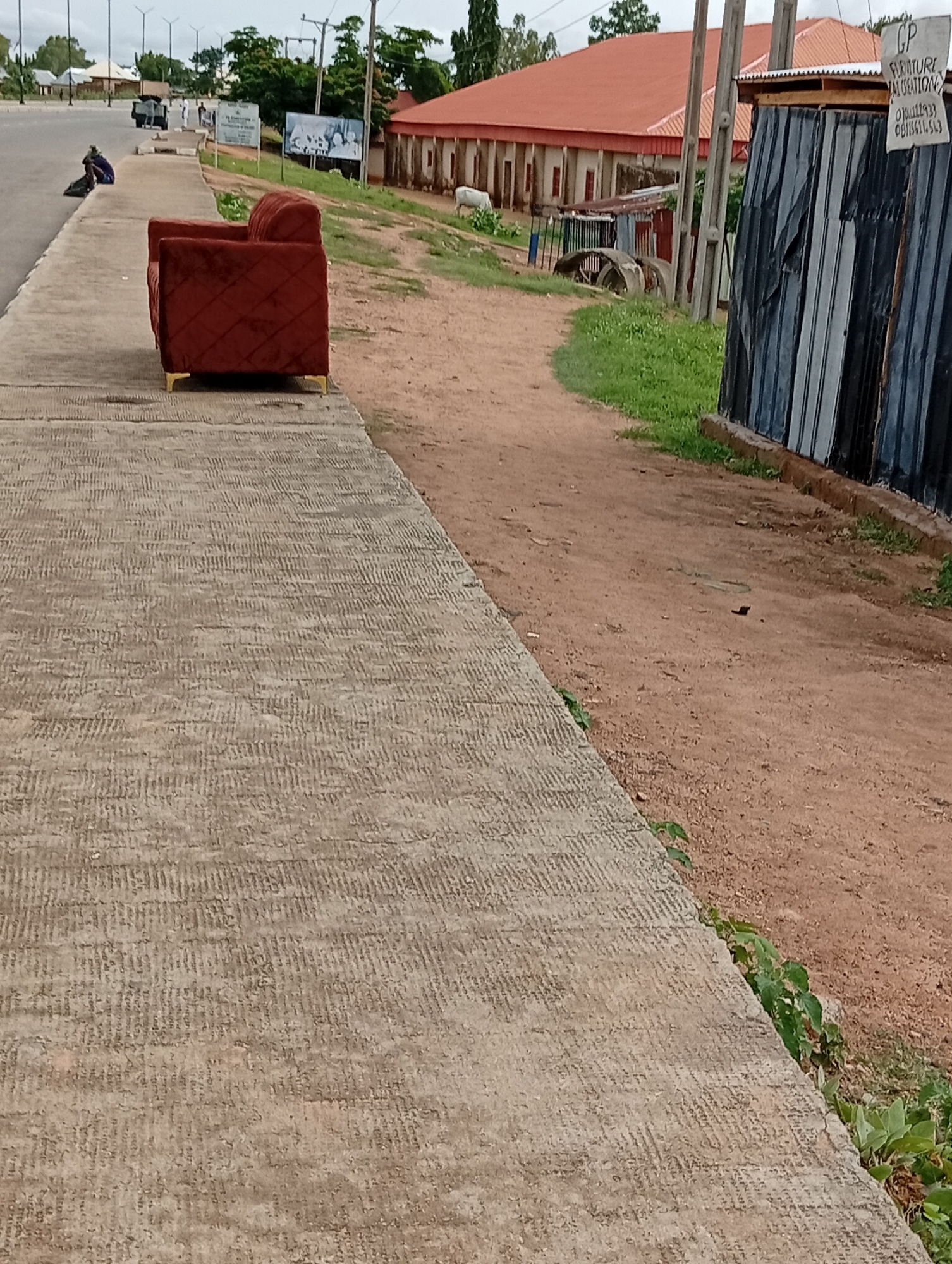 In Jalingo, Nigeria, a red upholstered sofa is placed on a concrete sidewalk adjacent to a dirt road. A person is seated on the ground near a black bag on the sidewalk, to the left of the sofa. In the background, a building with a red roof stands, and to its right, a corrugated metal structure displays a sign reading "CP FURNITURE CREATION" with a phone number. The environment appears to be an outdoor, possibly commercial area.