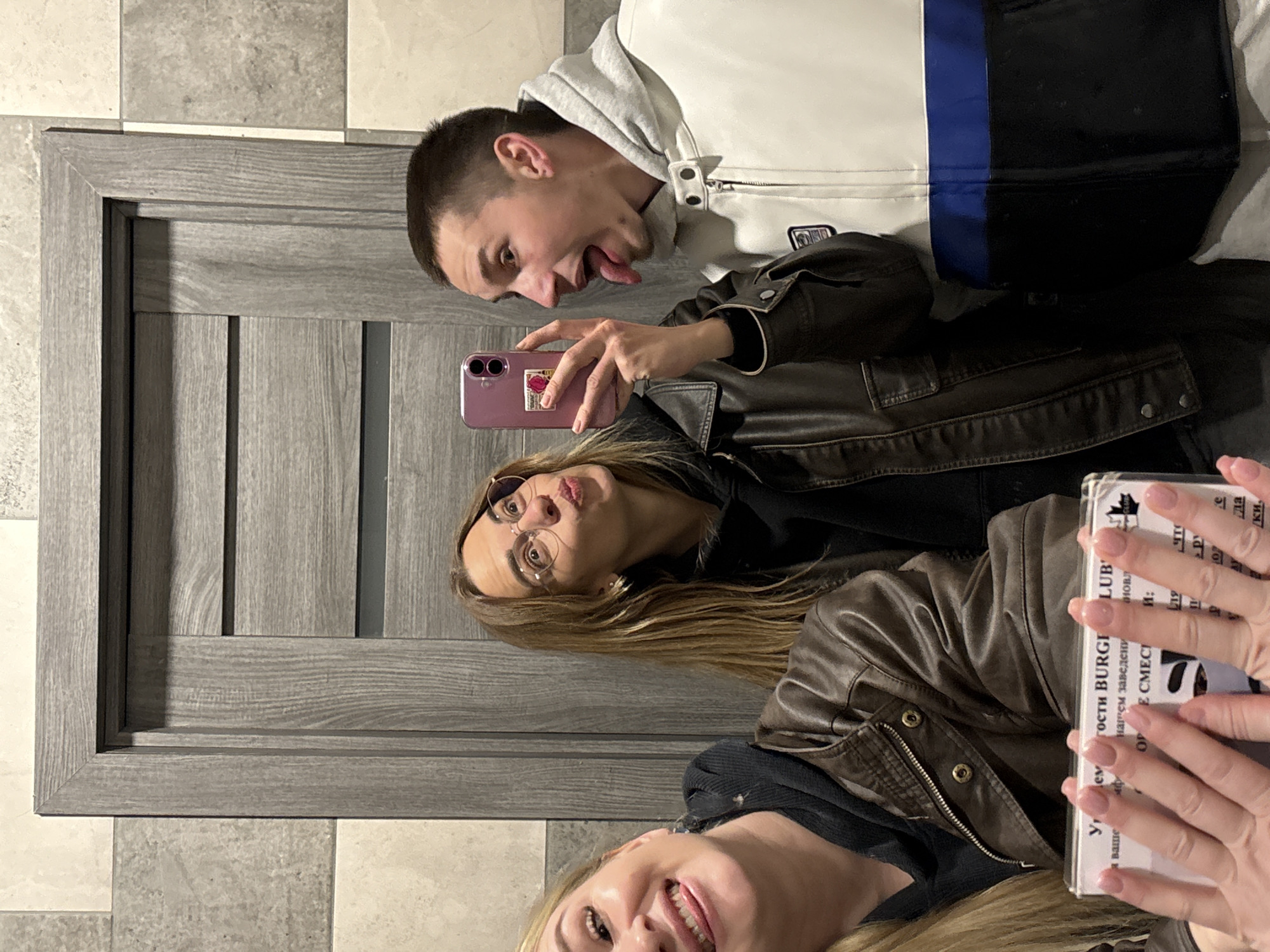 The image shows three people taking a selfie indoors. A man with short dark hair is at the top of the frame, sticking out his tongue. He is wearing a white, gray and black hooded jacket. Below him is a woman with long brown hair wearing glasses and a brown leather jacket, holding a pink phone. At the bottom of the frame, a woman with blonde hair is partially visible. Her hands are holding a book or document with text in Russian, including "BURGER CLUB". The background includes a gray door and checkered floor. It can be inferred that the location is Ust-Ilimsk, Russia due to the Russian text on the book.