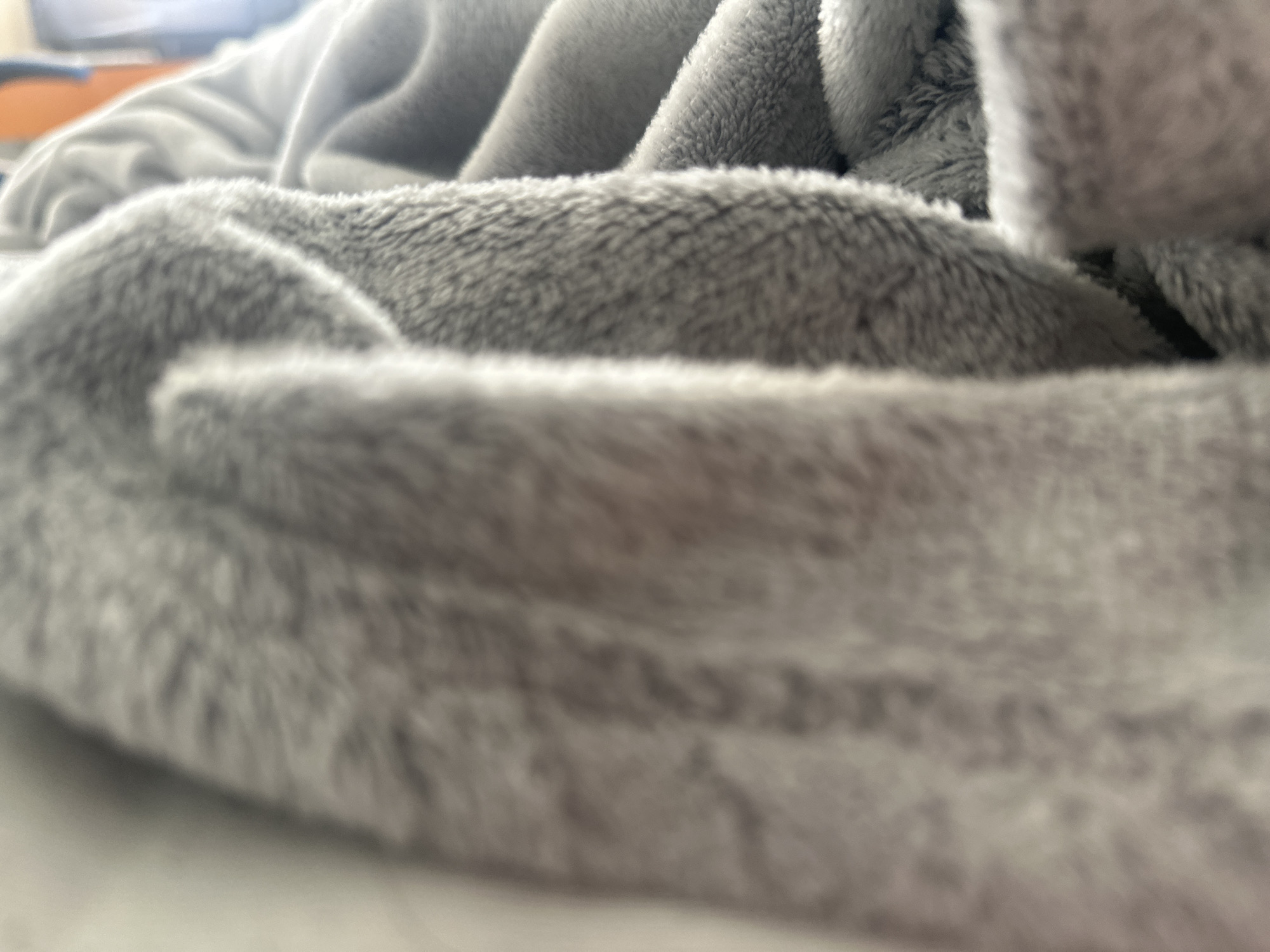 The image is a close-up, low-angle shot of a plush gray blanket. The blanket is folded and creased, creating soft shadows and highlights that emphasize its texture. The focus is shallow, with the foreground in sharp detail and the background blurred. The lighting appears soft and ambient. There is no discernible background or context to suggest a specific location in Shumen, Bulgaria, or any other city. The scene is devoid of people, actions, or interactions, offering only an abstract view of the fabric's texture and form.