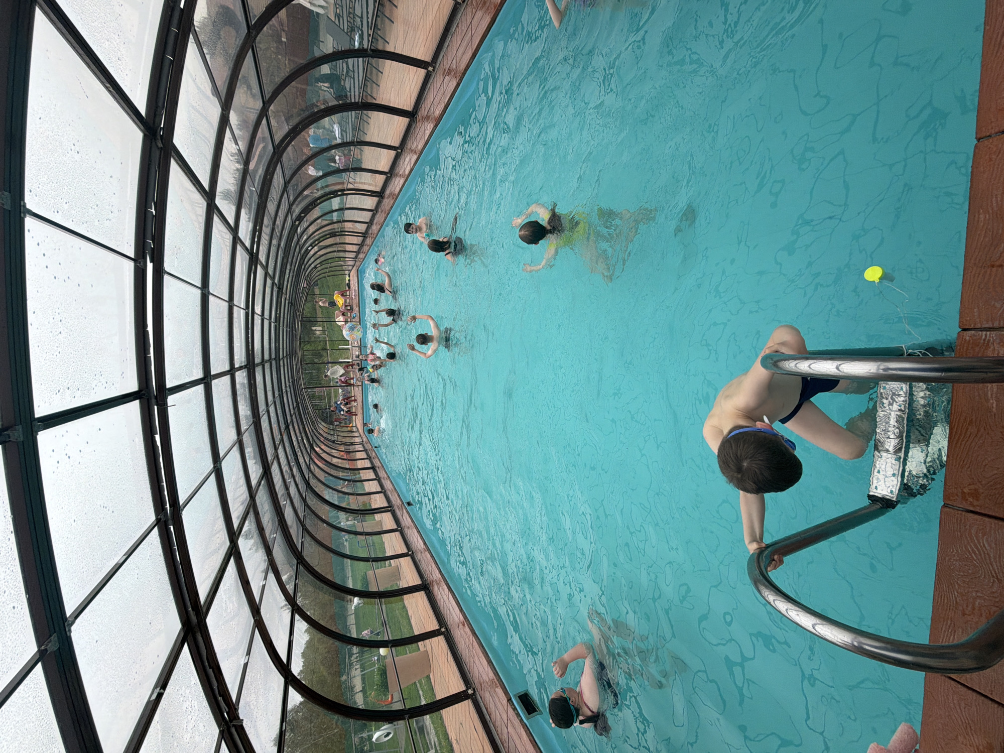 A high-angle, eye-level shot reveals an indoor swimming pool with a glass enclosure. The pool is filled with light blue water, in which several people are swimming or standing. A boy in dark blue swim trunks and blue goggles is seen holding onto a metal ladder, partially submerged. To the left, the glass enclosure extends, showing raindrops or condensation on the panels. Outside the enclosure, a glimpse of green grass and trees can be seen. The pool appears to be located in Birobidzhan, Russia.