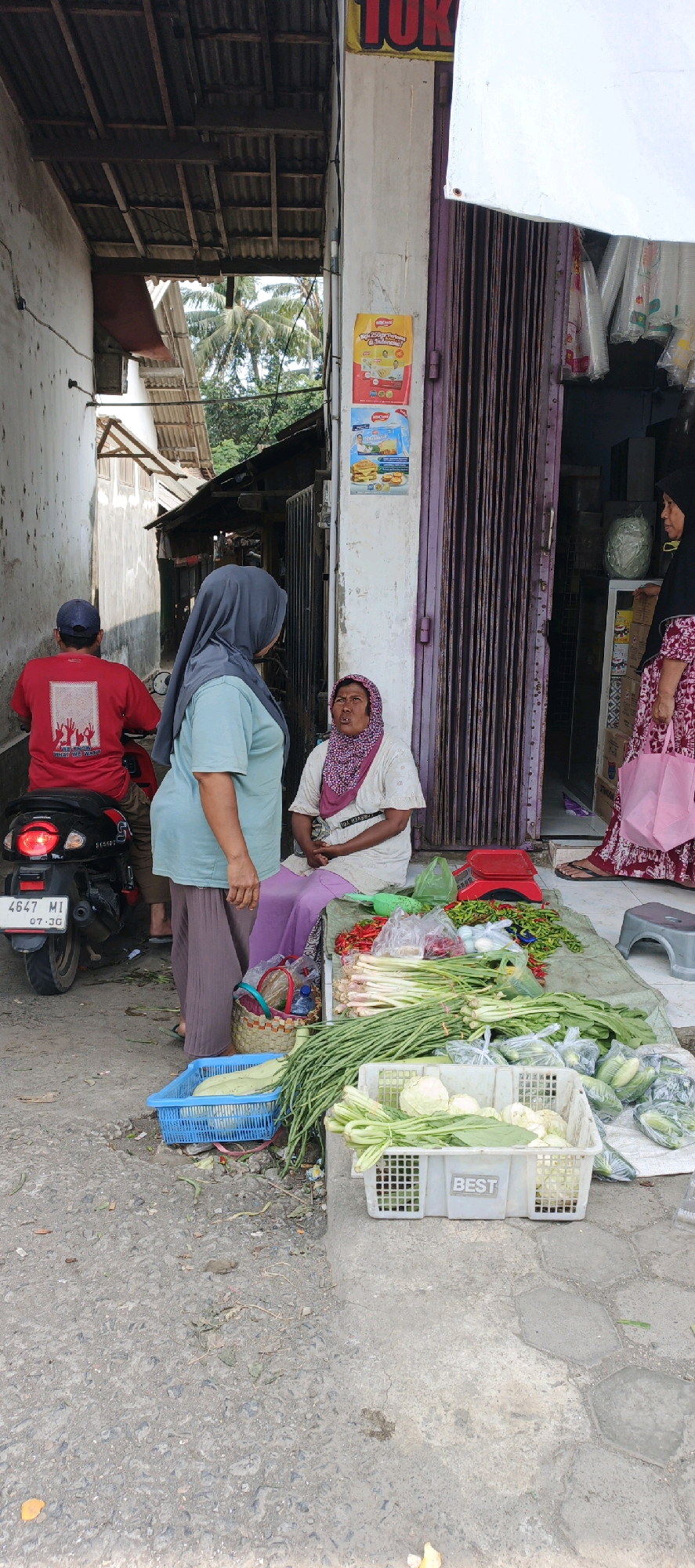 In Kasiyan, Indonesia, at an outdoor market, a woman wearing a patterned headscarf and a light-colored tunic sits on a purple cloth spread on the ground. She is looking upwards, her mouth slightly open. In front of her, a large selection of fresh vegetables is displayed on a green tarp. This includes bundles of long beans, stalks of lemongrass, leafy greens, and piles of red chilies. A red scale sits atop the tarp near the chilies. To her left, a woman in a gray hijab and a light blue shirt stands, facing away from the camera and towards the seated woman. To the left of these women, a person wearing a red shirt with a graphic design and a blue baseball cap is seated on a black motorcycle, also facing away from the camera. The motorcycle has a license plate that reads "4647 MI 07-36". The scene is set against a narrow alleyway with textured walls on the left and a shop entrance with purple corrugated doors on the right. Posters are visible on the wall to the right of the shop entrance. The ground is paved with a rough, grey surface.