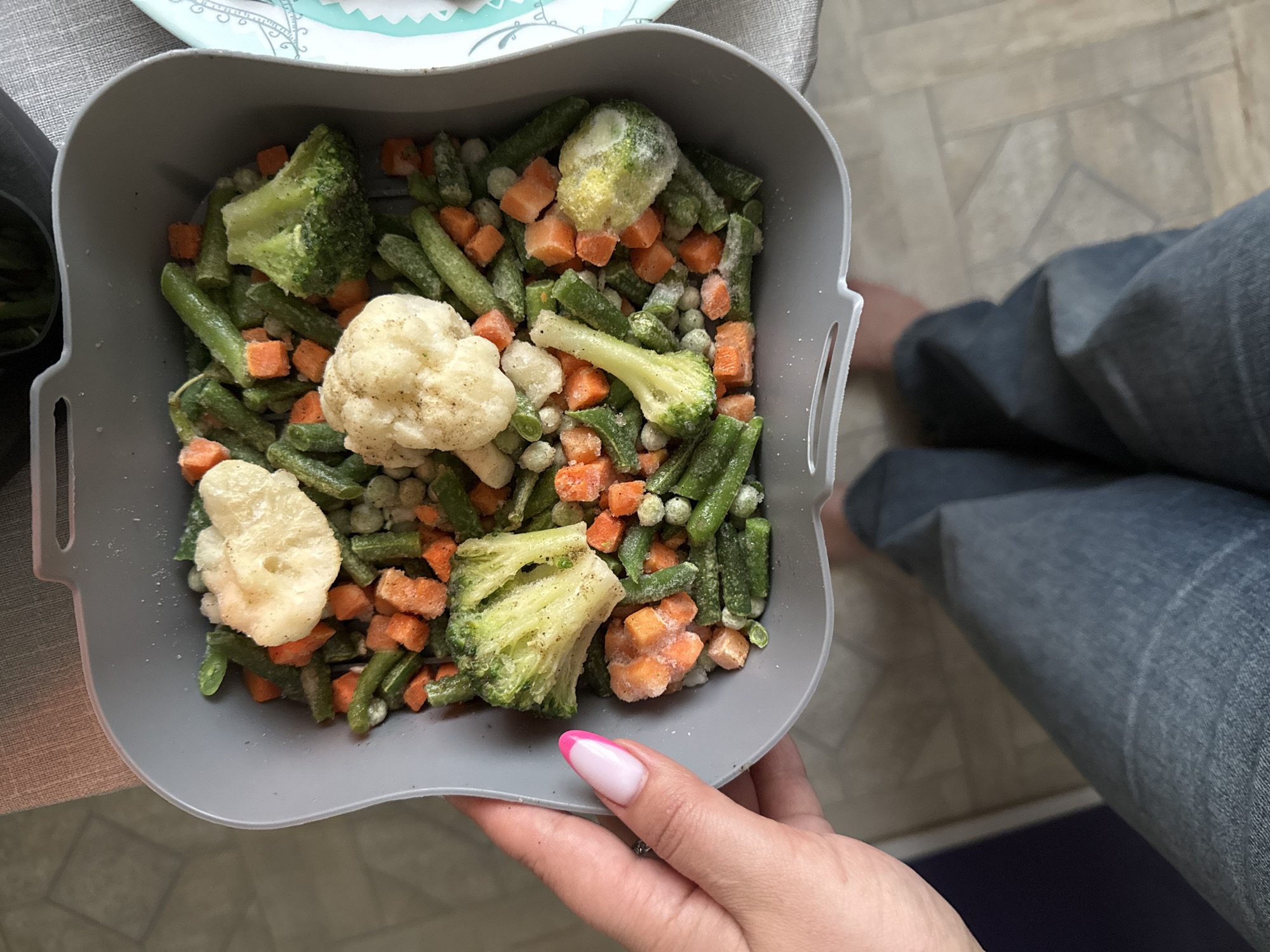 A person holds a gray square container filled with frozen mixed vegetables, including broccoli, cauliflower, carrots, green beans, and peas. The vegetables are visibly frosted. The person's hand, adorned with a pink-and-white manicured nail, supports the container. The person wears blue jeans. The background includes a countertop or table with a patterned surface, a partial view of another plate, and a glimpse of flooring. There is no visual information to confirm this image was taken in Chaikovsky, Russia.