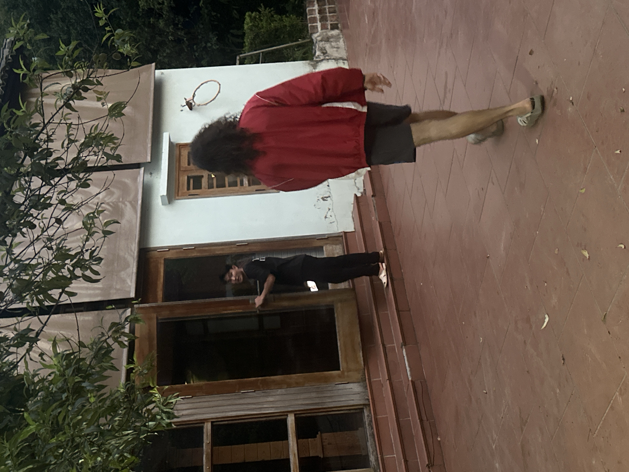The scene is outdoors, possibly in a residential area in Du Già Commune, Vietnam. The ground is paved with reddish-brown tiles. A white building is visible in the background, with large wooden-framed windows and doors. A person in black clothing is standing near the building, partially obscured by the open doorframe. Another person wearing a red jacket and dark shorts is walking on the paved area. A tree with green leaves is on the left side of the image. A metal ring, possibly part of a basketball hoop, is attached to the wall of the building.