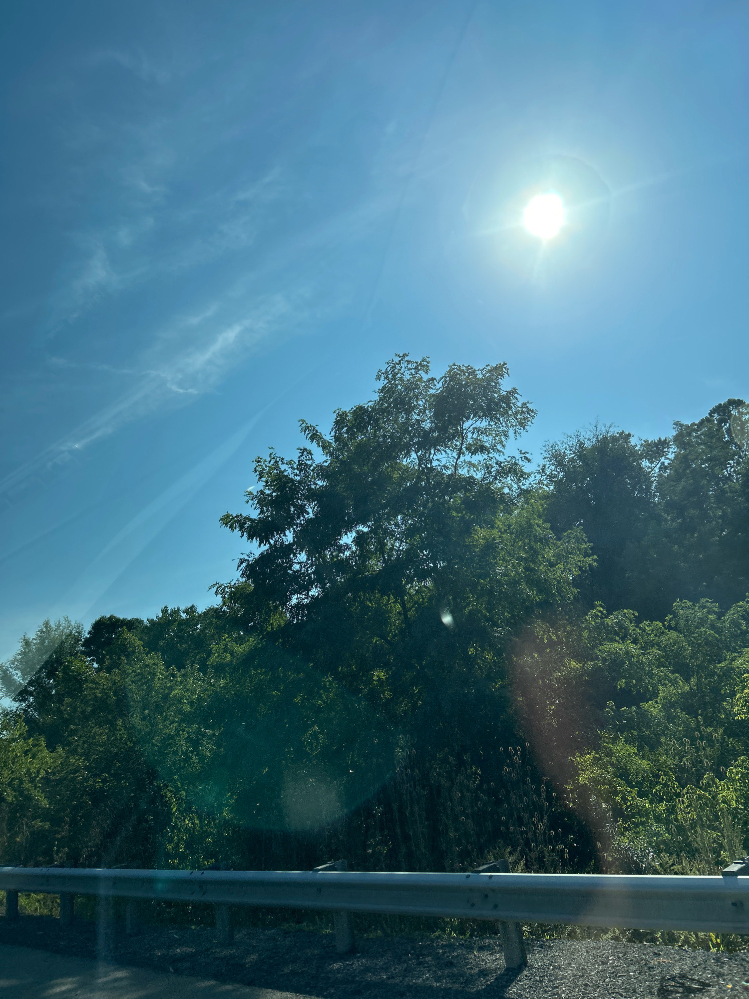 The image depicts a bright, sunny day in a rural or suburban area, likely Scott Township, United States of America. The sun is prominently visible in the upper right quadrant, casting a strong glare and creating lens flares across the image. A clear blue sky with wispy white clouds stretches across the top portion. Dense green foliage, consisting of trees and bushes, dominates the middle and lower sections of the frame, suggesting a natural or park-like setting. In the foreground, a silver metal guardrail runs horizontally, indicating the presence of a road or pathway. The ground beneath the guardrail appears to be gravel or dirt. No people or animals are visible in the scene. The overall impression is one of a serene, outdoor environment bathed in sunlight.