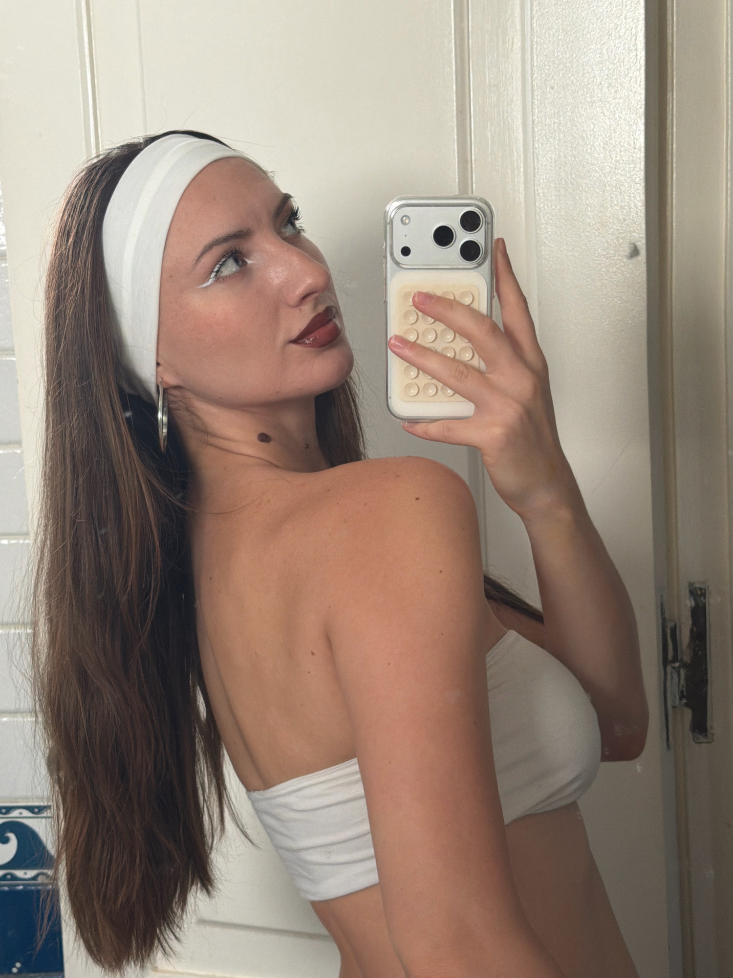In a bathroom with white tiled walls and a white door, a person with long brown hair pulled back by a white headband and wearing a white strapless top is taking a selfie in a mirror. Their makeup includes dramatic white eyeliner and dark lipstick. They are wearing a large silver hoop earring and have a mole on their neck. The person is holding a clear phone case with a cream-colored pop socket on the back. The scene is devoid of any other people or identifiable objects related to Sharm El Sheikh, Egypt.