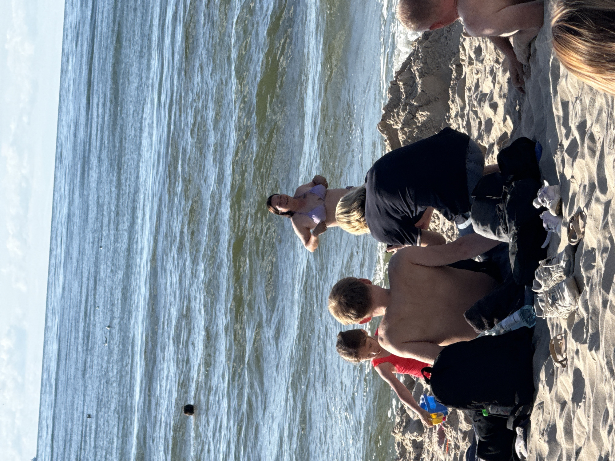 The image depicts a sunny day at the beach in Międzyzdroje, Poland. Several people are visible on the sandy shore, with the ocean and waves in the background. A woman in a purple bikini is lying on her side near the water's edge, smiling. Closer to the shore, a group of people, including children, are gathered. Some are partially submerged in the water, while others are on the sand. One child is wearing a red top and appears to be holding a toy. Objects like a backpack and shoes are also scattered on the sand. The scene suggests a relaxed day of recreation and enjoyment at the beach. The bright sunlight and the presence of people in swimwear indicate warm weather and a typical summer day.