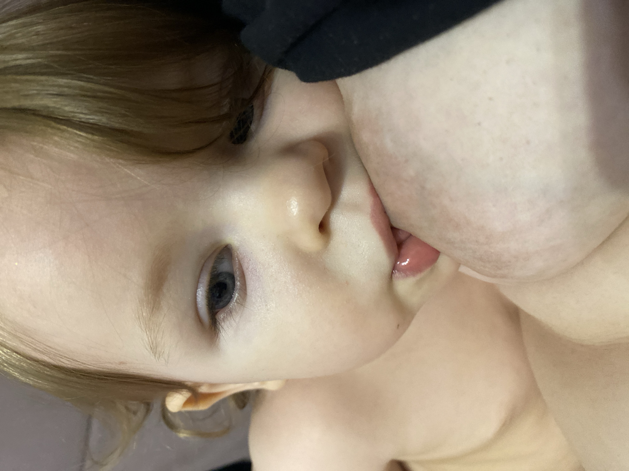 A close-up image shows a fair-skinned child, likely a toddler, with light blonde hair and blue eyes. The child is in close proximity to a person, with their face near the person's breast. The child's mouth is slightly open and possibly latched onto the breast. The person's skin tone is similar to the child's. The child's body is partially visible, and the environment is out of focus. The location of the scene is unknown.