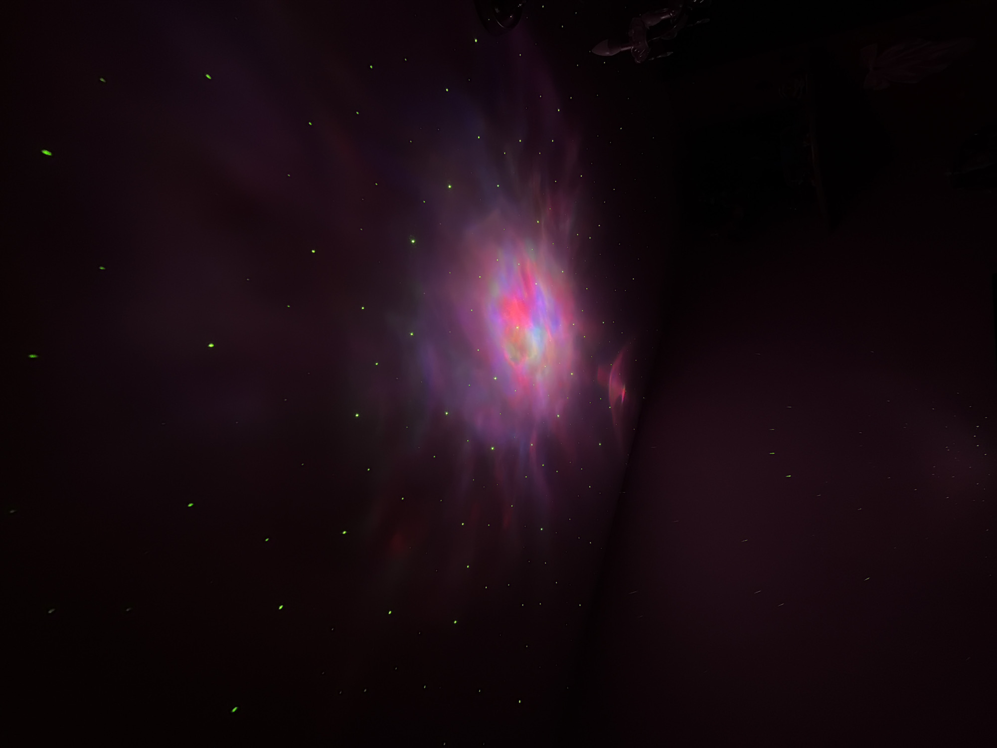 The image shows a dark room with a projected nebula-like display on the ceiling. The display is a mix of purple, pink, red, and blue hues, suggesting a cosmic or fantasy-themed lighting effect. Small green dots are scattered throughout the dark areas, likely representing stars. The room appears to have a wall on the right side, and the overall ambience is dim and theatrical. The location in the city of Perm, Russia is not explicitly evident in this image, so that location is unknown.