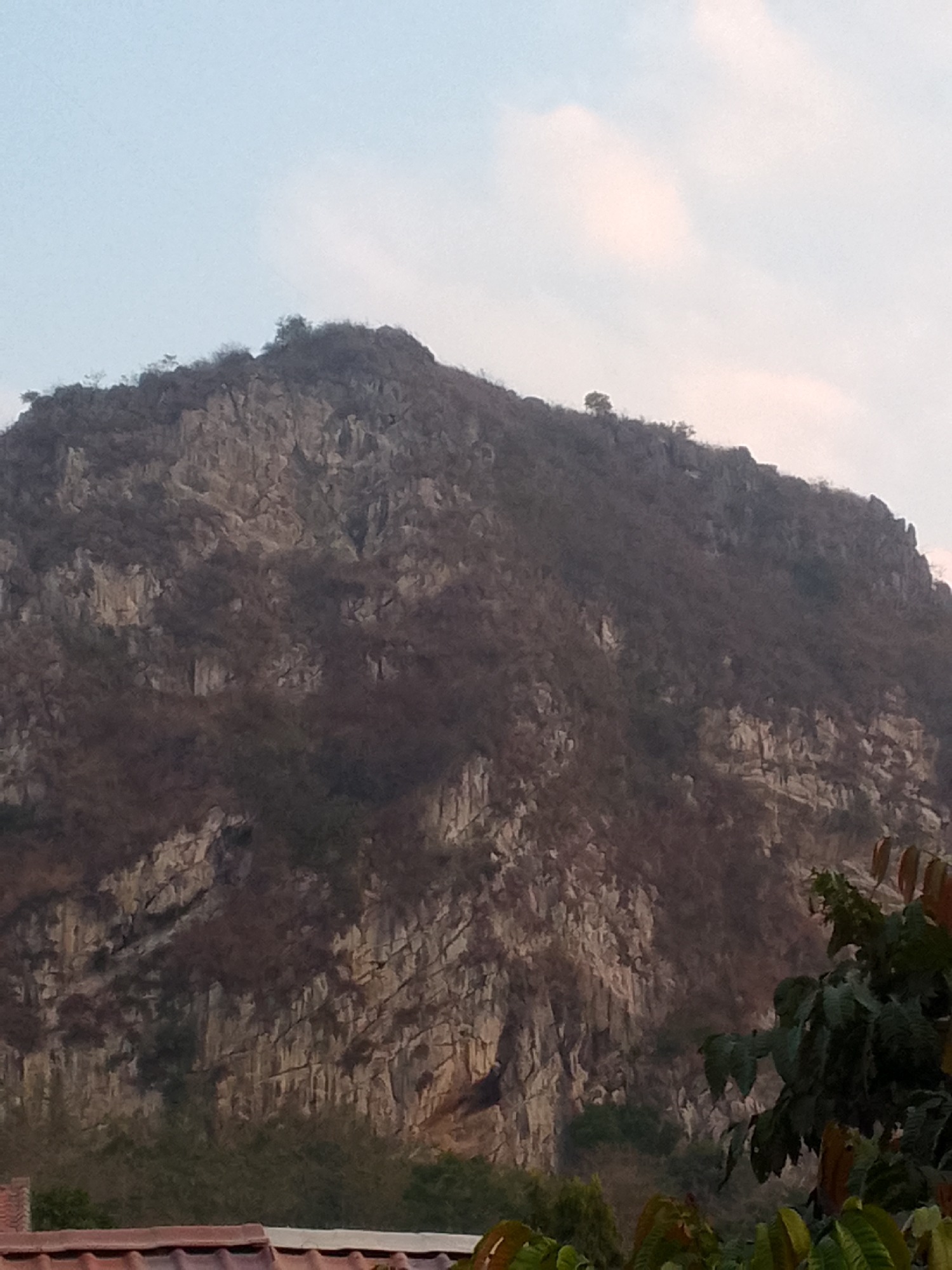 The image shows a mountain in Gunungmasigit, Indonesia. The mountain is rocky and covered in sparse vegetation. The sky is visible in the background, with some clouds. In the foreground, there is a portion of a tiled roof, indicating the presence of a building. Some green foliage is also visible in the lower right corner of the image. The scene appears to be a natural landscape with hints of human habitation.
