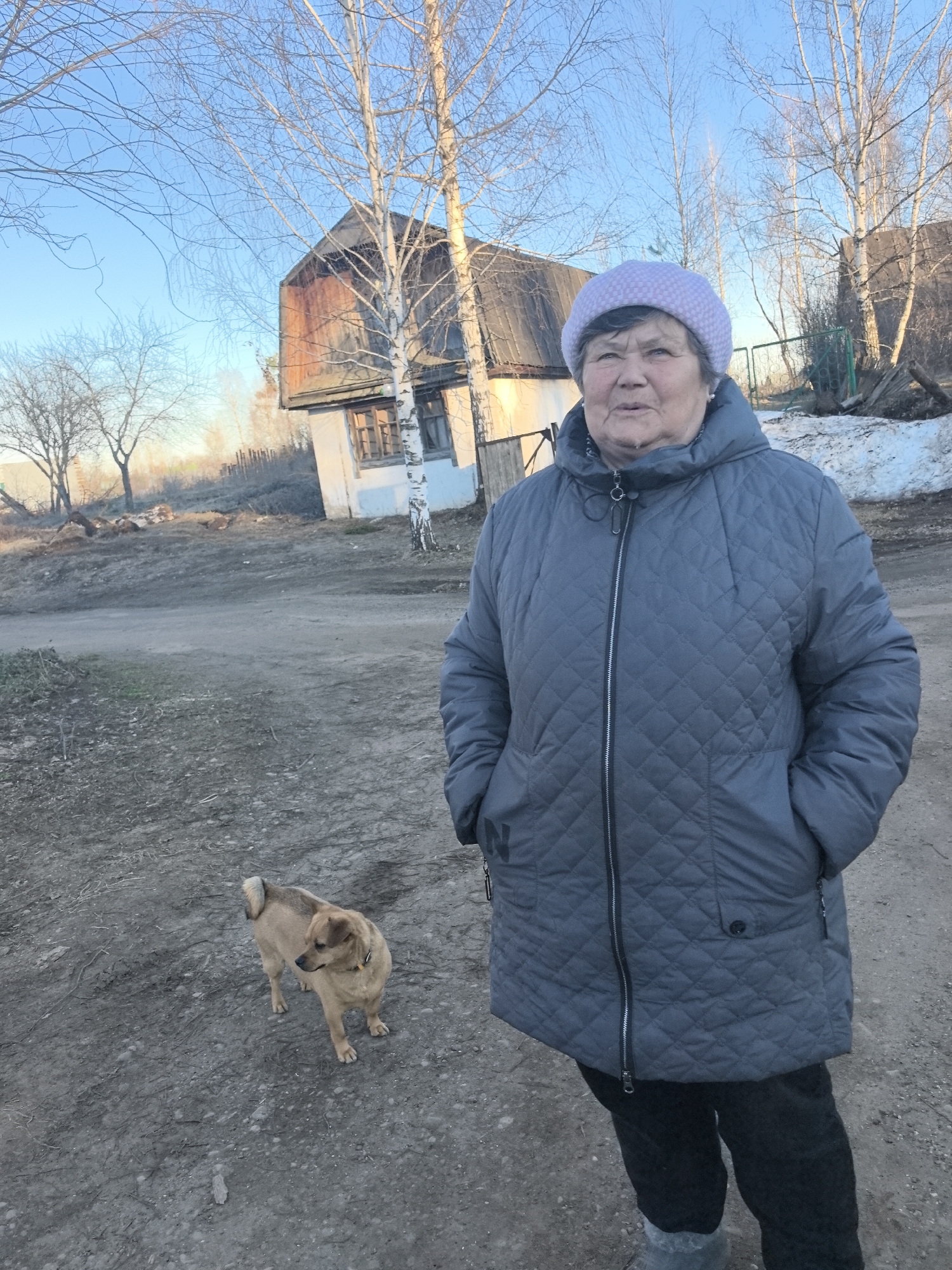 The image shows an older woman wearing a quilted grey coat and a light purple hat. She stands on a dirt road with her hands in her pockets. A small, tan dog is near her feet. Behind her is a small, weathered wooden house with bare birch trees surrounding it. A patch of snow can be seen near the house. The location is in the city of Noviy, Russia.
