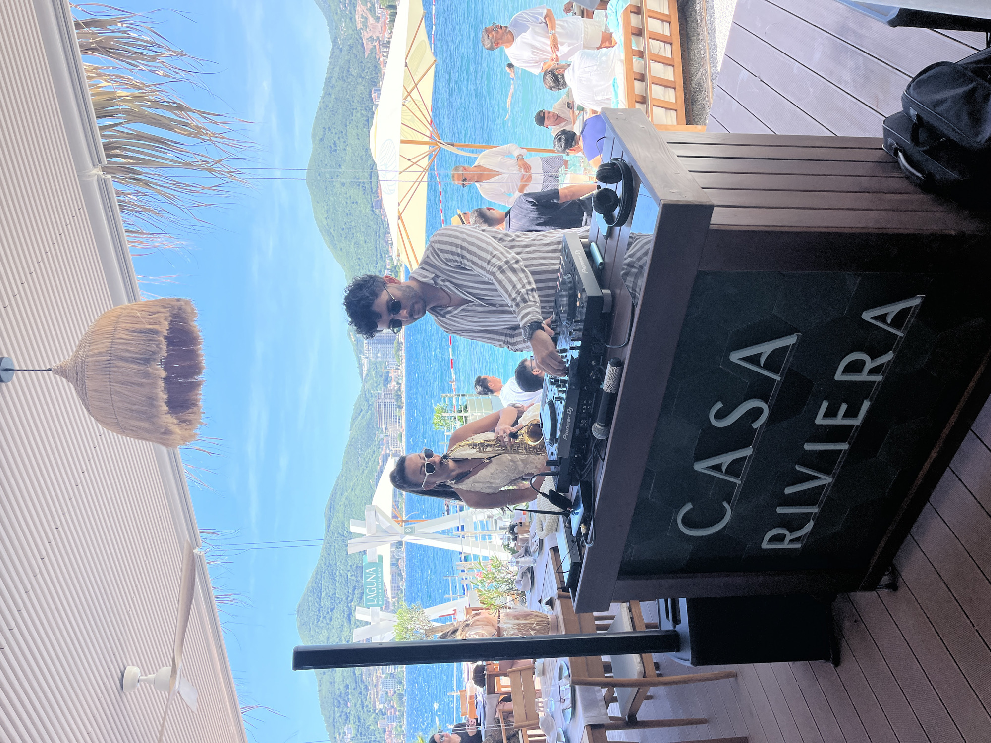 The image depicts a lively scene at Casa Riviera Beach Restaurant in Budva, Montenegro. In the foreground, a DJ booth is set up, complete with a Pioneer DJ controller and headphones. A male DJ, wearing a striped shirt and sunglasses, is actively engaged in his work. To his left, a woman in a white dress and sunglasses plays the saxophone, adding a musical element to the atmosphere.
The background reveals the restaurant's outdoor seating area, with tables and chairs set for dining. Beyond the restaurant, the turquoise waters of the Adriatic Sea are visible, with several people enjoying the beach and swimming. Hills covered in greenery and buildings rise in the distance, characteristic of Budva's scenic coastal landscape. The overall ambiance is one of a relaxed and enjoyable beachfront dining and entertainment experience.