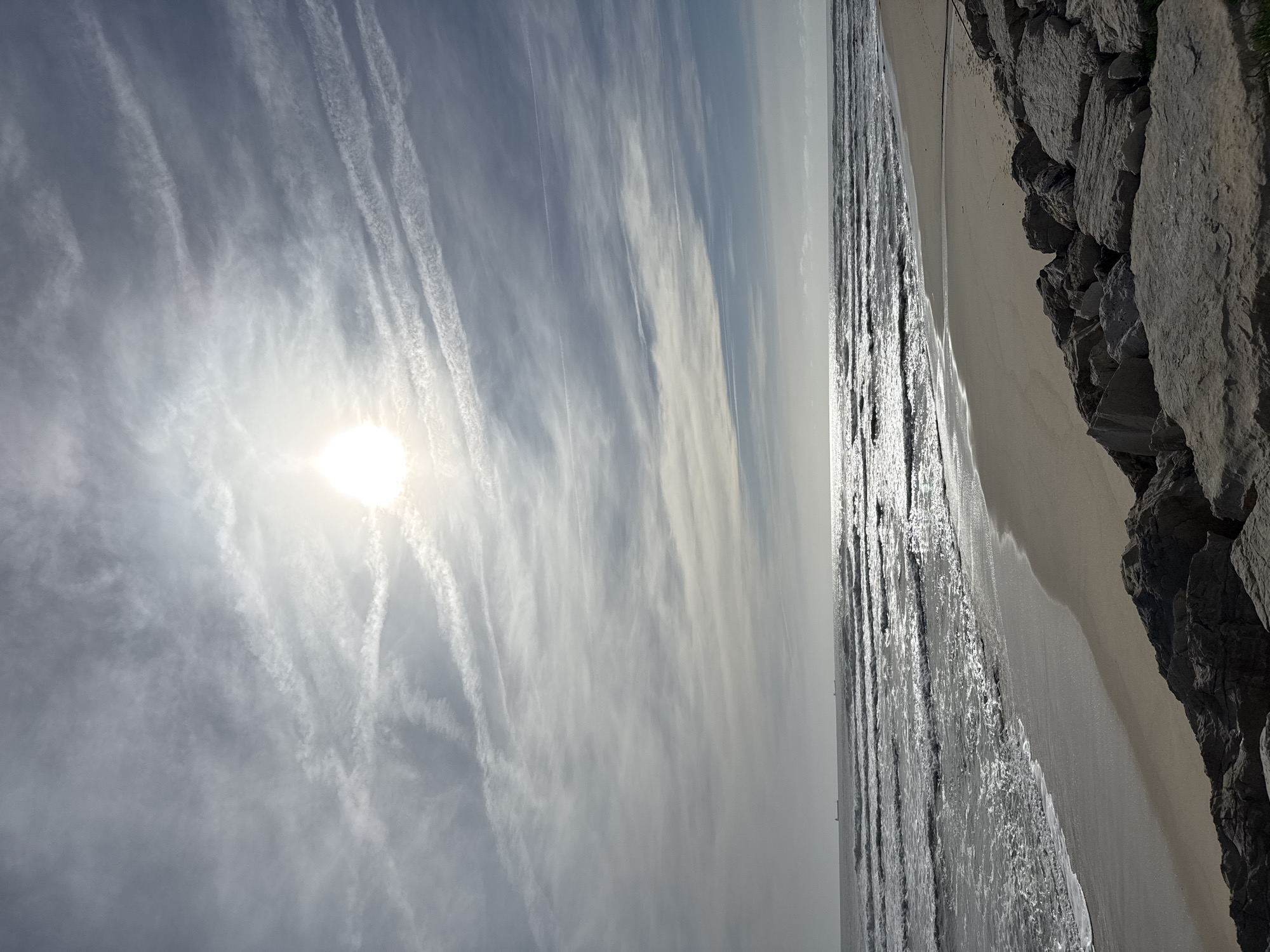 The image displays a sunny coastal landscape in Figueira da Foz, Portugal. The scene is dominated by a bright sky, an expansive ocean, a sandy beach, and a rocky embankment.
In the upper-left quadrant, the sun shines intensely, creating a strong glare and scattered light through the clouds. The sky is largely blue, adorned with numerous long, wispy white clouds, some appearing as distinct linear contrails stretching across the frame.
Below the horizon, the ocean occupies the middle and lower sections of the image. Its surface reflects the bright sunlight, creating shimmering, elongated streaks of light, particularly where gentle waves break near the shore. A strip of light-colored sandy beach runs along the water's edge, creating a smooth transition from the ocean.
On the right side of the image, a formation of large, dark grey, irregular rocks forms a boundary, suggesting a natural cliff face or a man-made breakwater.
There are no people or specific activities depicted. The time of day appears to be either morning or late afternoon due to the strong, direct sunlight and reflections. The weather is clear and bright, with high-altitude clouds. No text is visible in the image. The overall impression is one of natural beauty and tranquility.